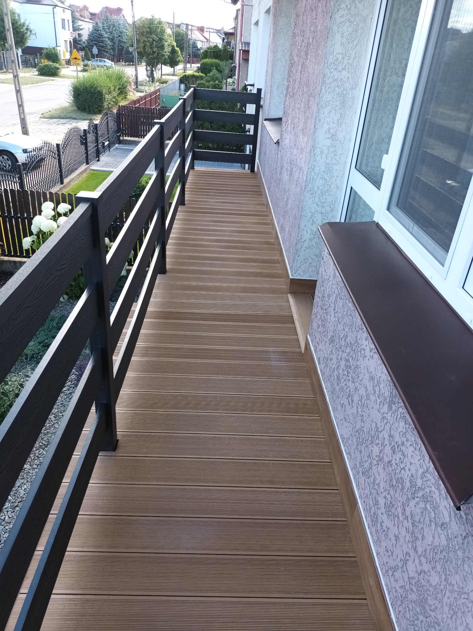 Długi, wąski balkon z brązową, kompozytową podłogą imitującą drewno, oraz ciemnoszarą, nowoczesną balustradą. Widok na ulicę z zabudową jednorodzinną i ogrodami.