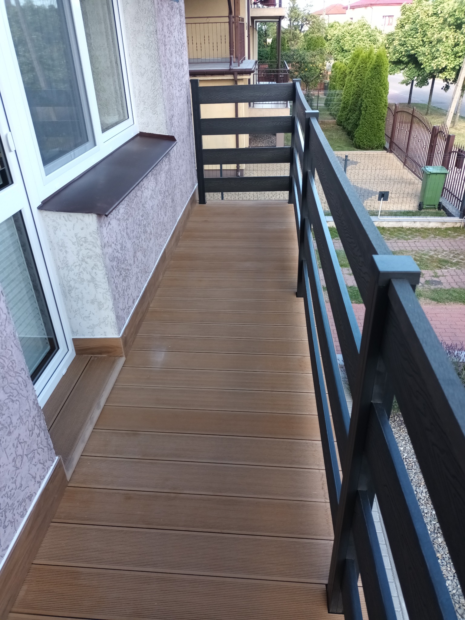 Zadaszony balkon z podłogą z desek kompozytowych w odcieniach brązu i czarną, drewnianą balustradą; widok na posesję z zielonym trawnikiem i ogrodzeniem.