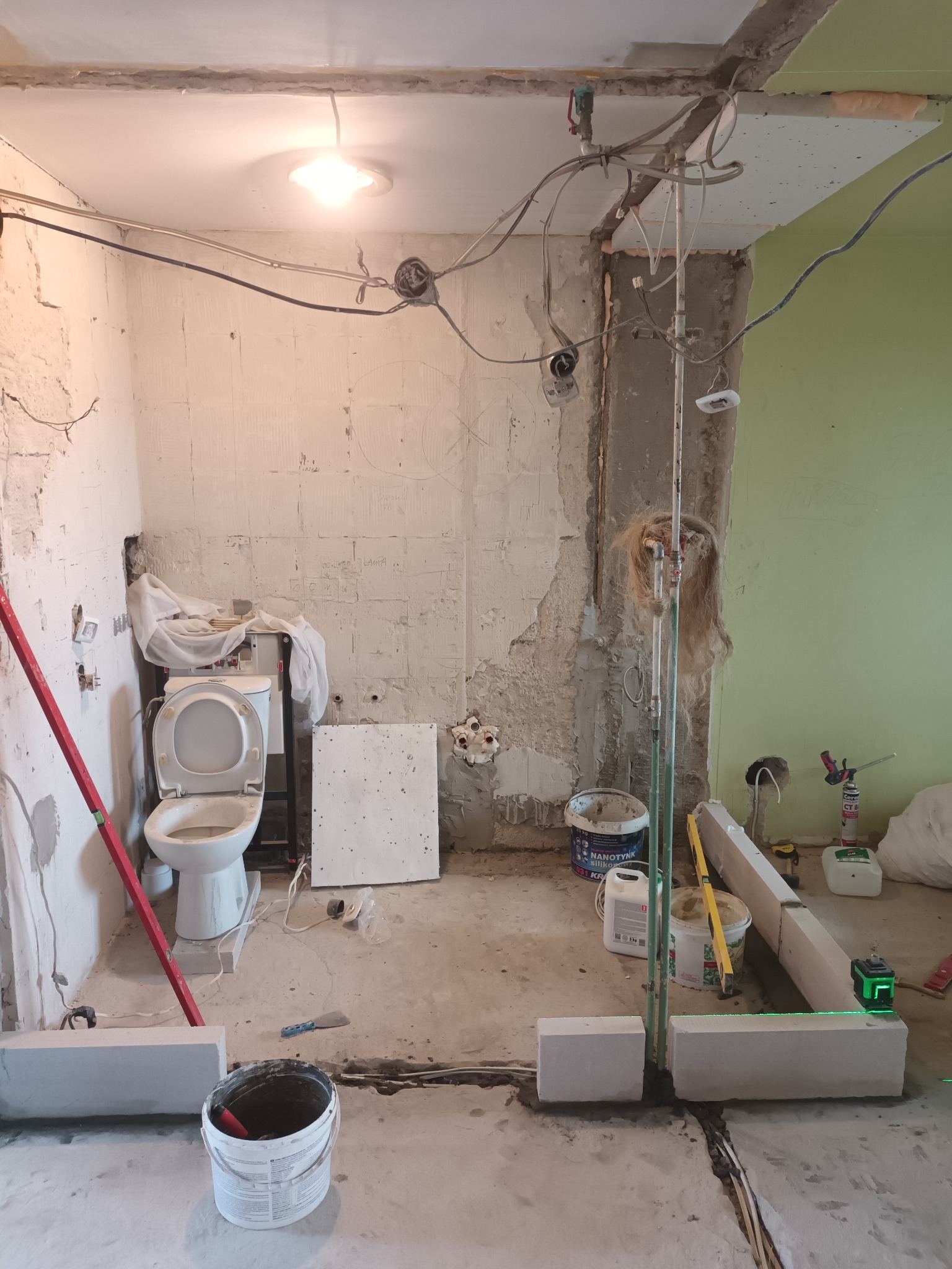 Wnętrze łazienki w trakcie prac wykończeniowych, widoczne odsłonięte instalacje, surowa ściana z resztkami tynku, częściowo pomalowana na zielono. Ustawiona toaleta, wiadro z zaprawą, poziomica...