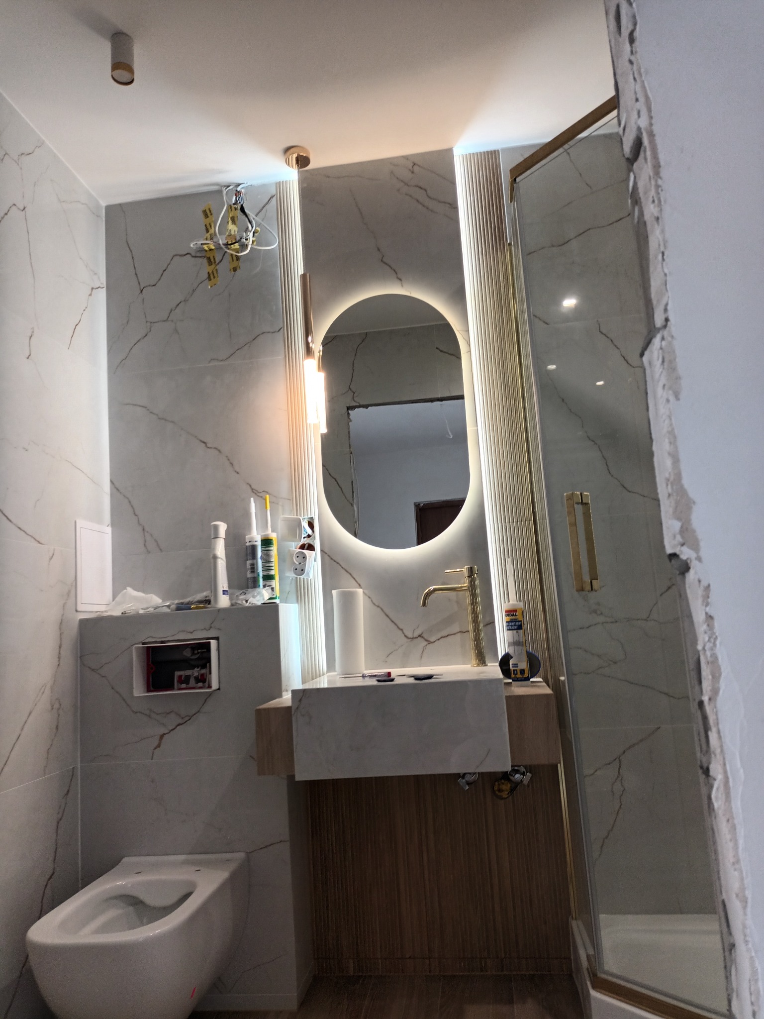 Wnętrze łazienki w trakcie prac wykończeniowych: częściowo zamontowana umywalka z szafką, toaleta, lustro z podświetleniem LED, widoczne narzędzia i materiały budowlane, ściany wyłożone płytkami...