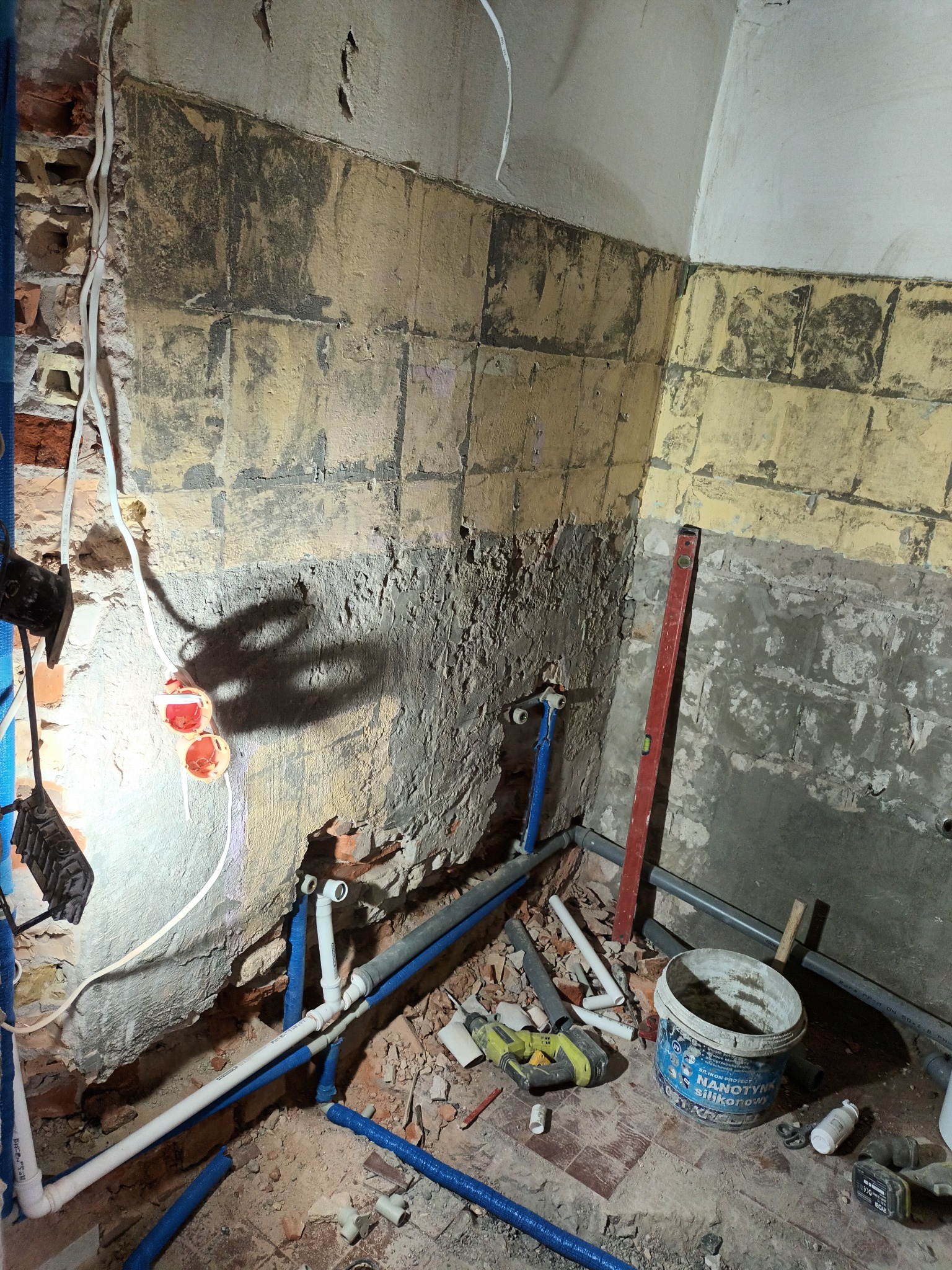 Łazienka w trakcie remontu: odkryte instalacje wodne, ściany z częściowo usuniętymi płytkami, poziomica oparta o ścianę, wiadro z silikonem, narzędzia leżące na podłodze.