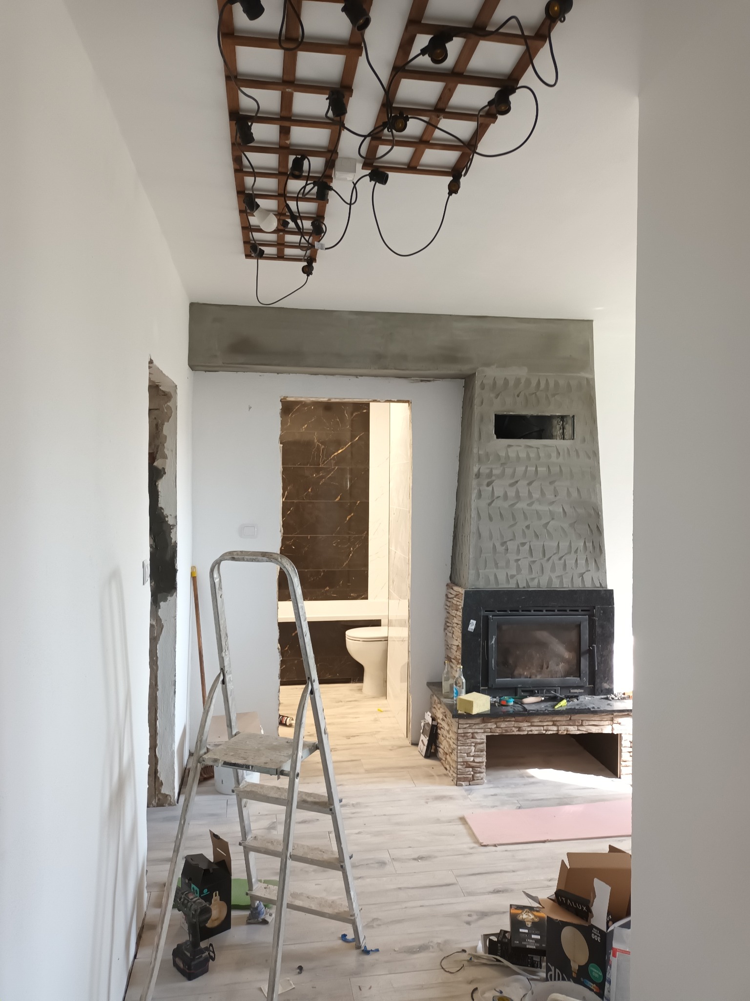 Wnętrze w trakcie prac wykończeniowych: widoczna drabina, surowy kominek z fakturą, instalacja oświetleniowa z drewnianą kratownicą i wiszącymi żarówkami, oraz fragment łazienki z ciemnymi płytkami...