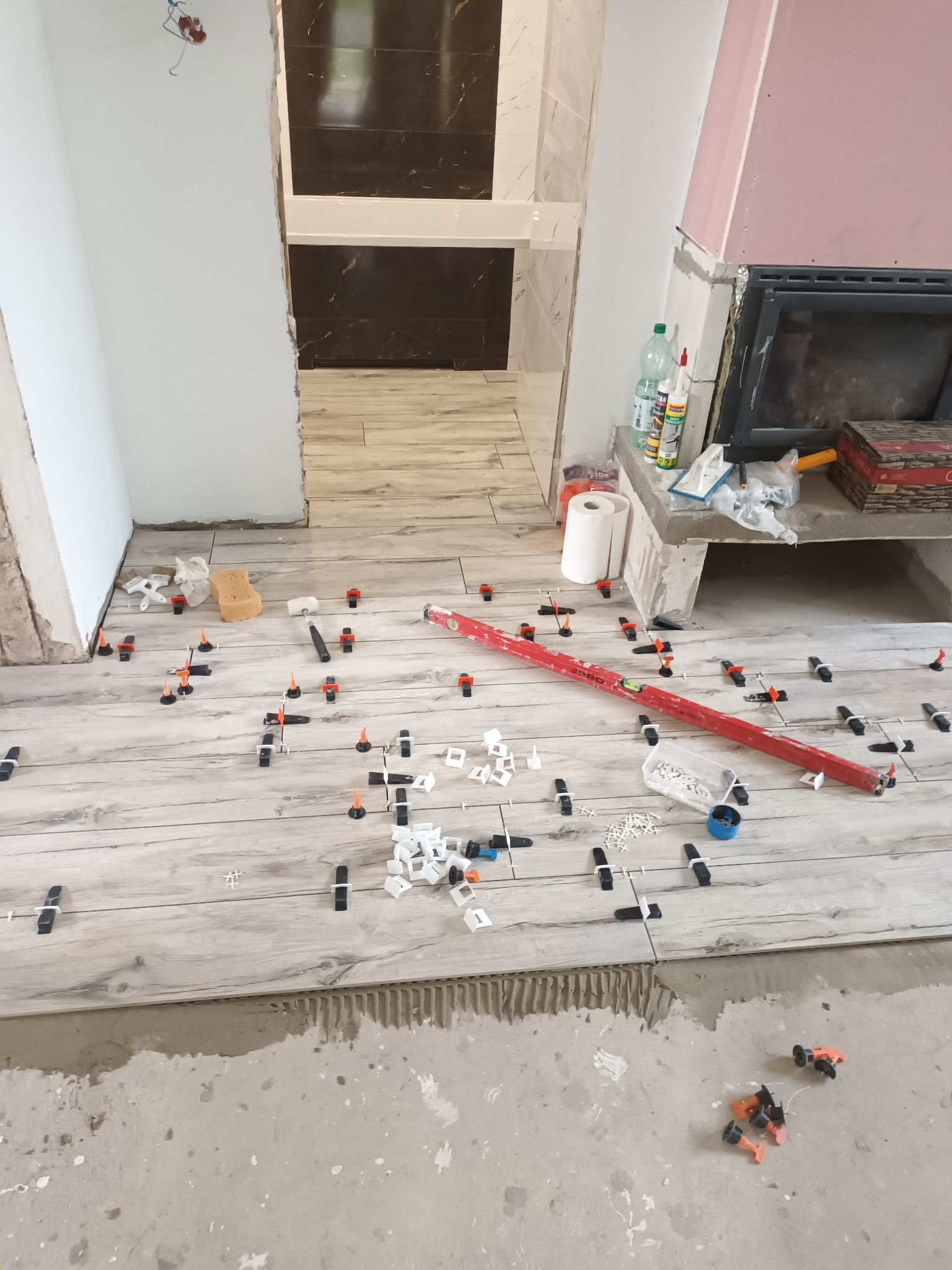 Podłoga w trakcie układania płytek imitujących drewno, widoczne poziomica, krzyżyki dystansowe, klej i narzędzia glazurnicze, w tle fragment łazienki z ciemnymi płytkami na ścianie i obudowa kominka.