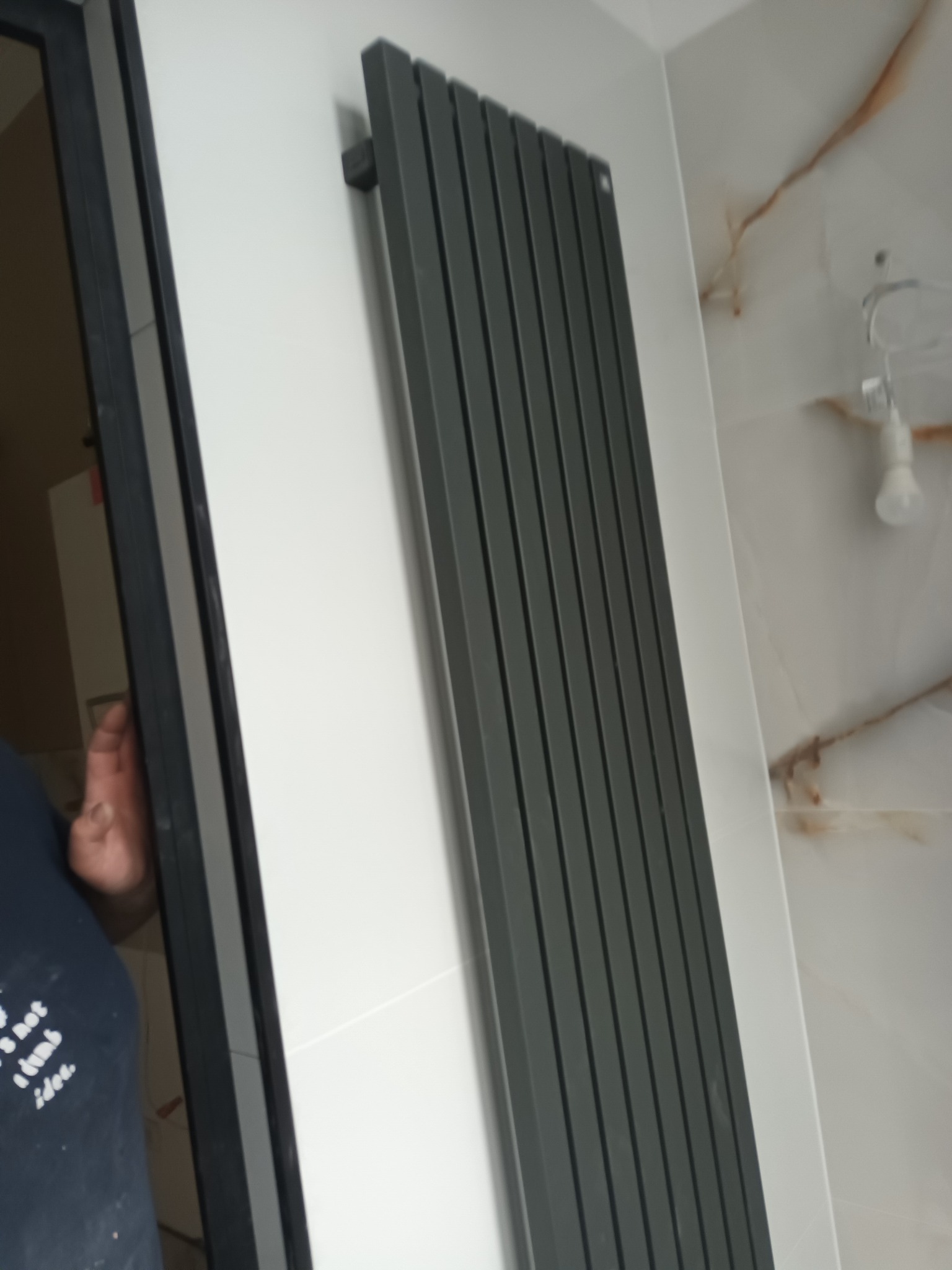 Szary, pionowy grzejnik panelowy zamontowany na białej ścianie z marmurowym wzorem w wykończonej łazience, widoczna osoba w tle.