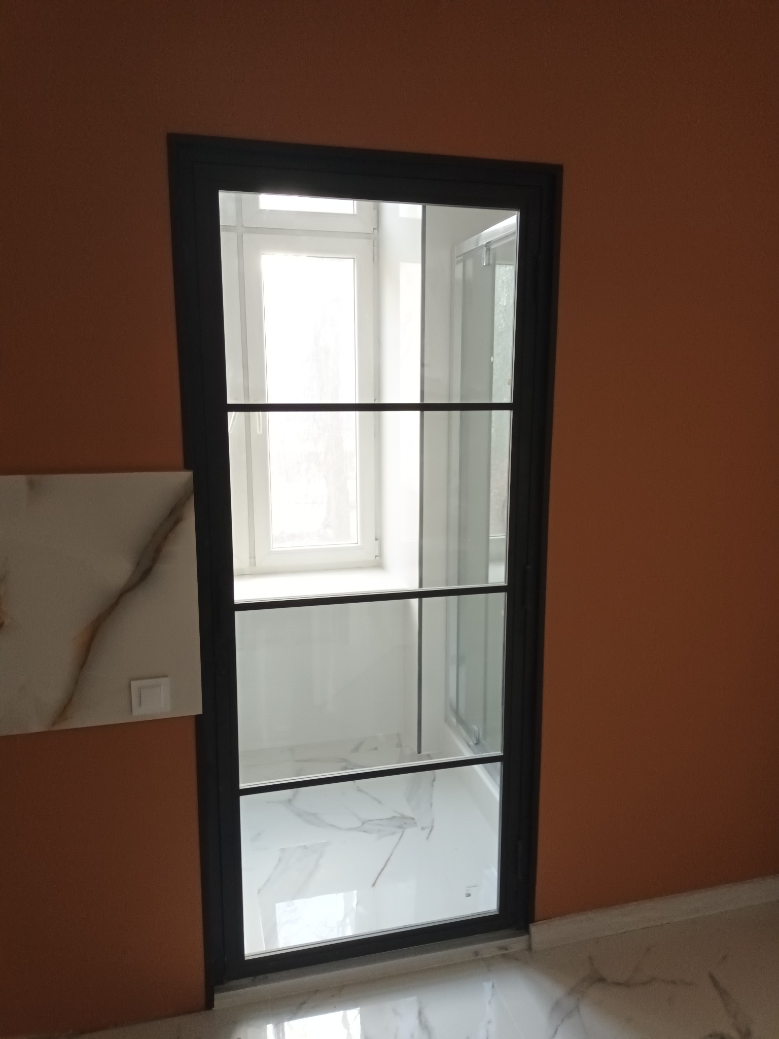 Przeszklone drzwi z czarnymi szprosami oddzielające przestrzeń z oknem od łazienki z kabiną prysznicową i marmurową podłogą, widoczne z perspektywy pomarańczowej ściany z fragmentem marmurowej...