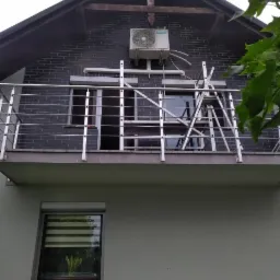 Zewnętrzna jednostka klimatyzacji zamontowana na elewacji budynku z czarnej cegły, widoczna drabina i barierka balkonu.