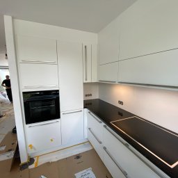 Gregor Möbel - Montaż białej kuchni z czarnym blatem i płytą indukcyjną, widoczne kartony zabezpieczające podłogę i odkurzacz przemysłowy, osoba w tle.