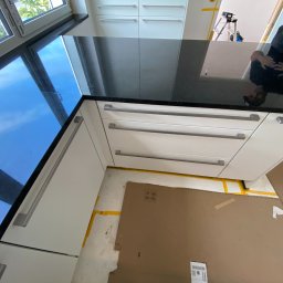 Gregor Möbel - Czarny blat kuchenny z widocznym odbiciem osoby, białe szafki z poziomymi uchwytami, okno z widokiem na zieleń na zewnątrz, podłoga zabezpieczona kartonem.