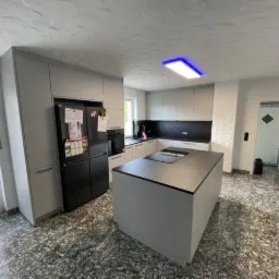 Nowoczesna kuchnia w szarościach z wyspą i czarnym sprzętem AGD, widok na nieregularną podłogę i drzwi wewnętrzne.