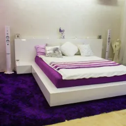 Nowoczesna sypialnia z białym łóżkiem na platformie, fioletowym materacem i puszystym dywanem w kolorze fuksji, uzupełniona o białe głośniki podłogowe i dekoracyjną figurkę anioła.