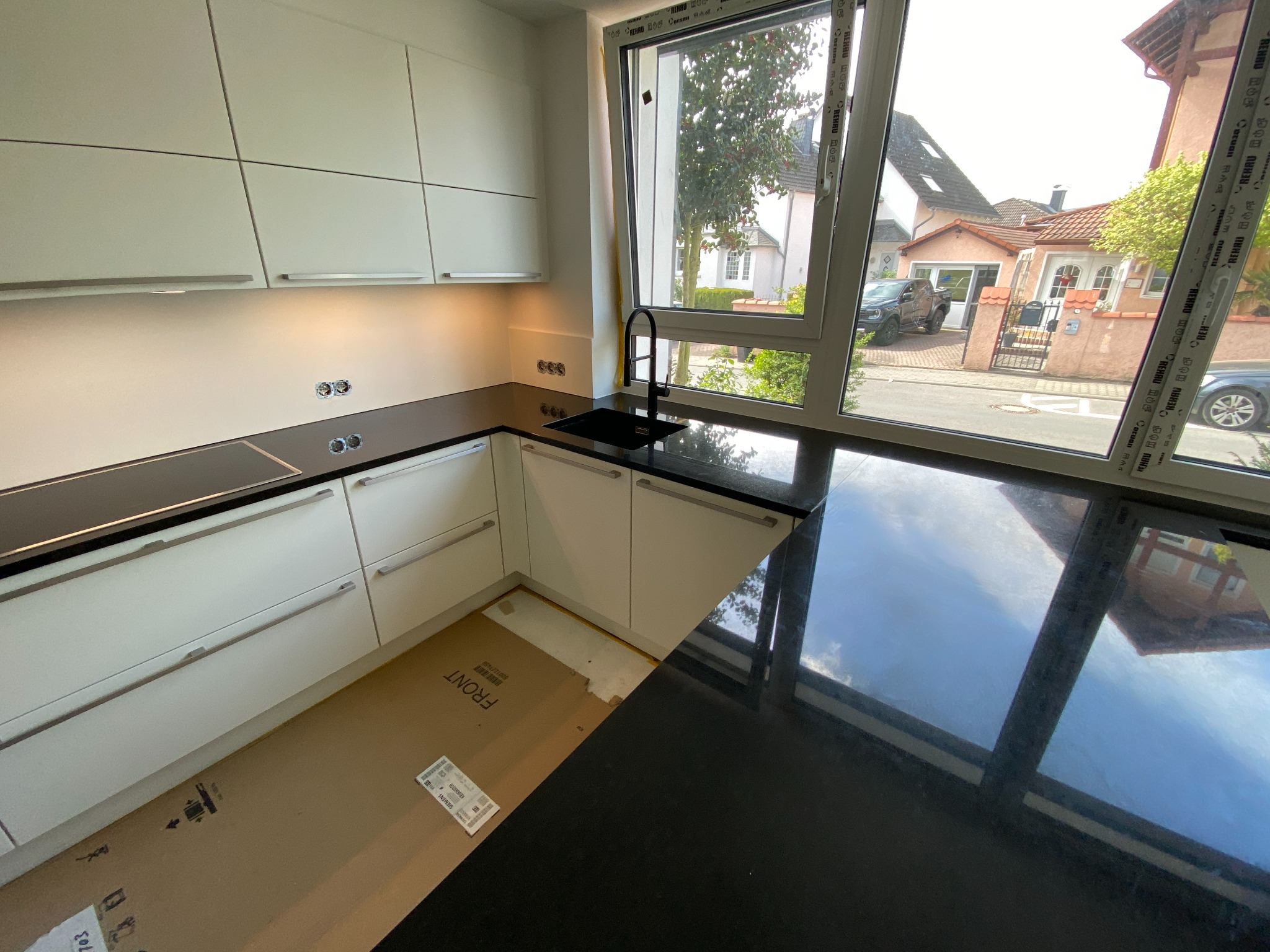 Wnętrze kuchni w trakcie montażu: białe szafki z poziomymi uchwytami, czarny blat i zlewozmywak przy oknie z widokiem na dom. Na podłodze kartonowe opakowania po elementach meblowych.