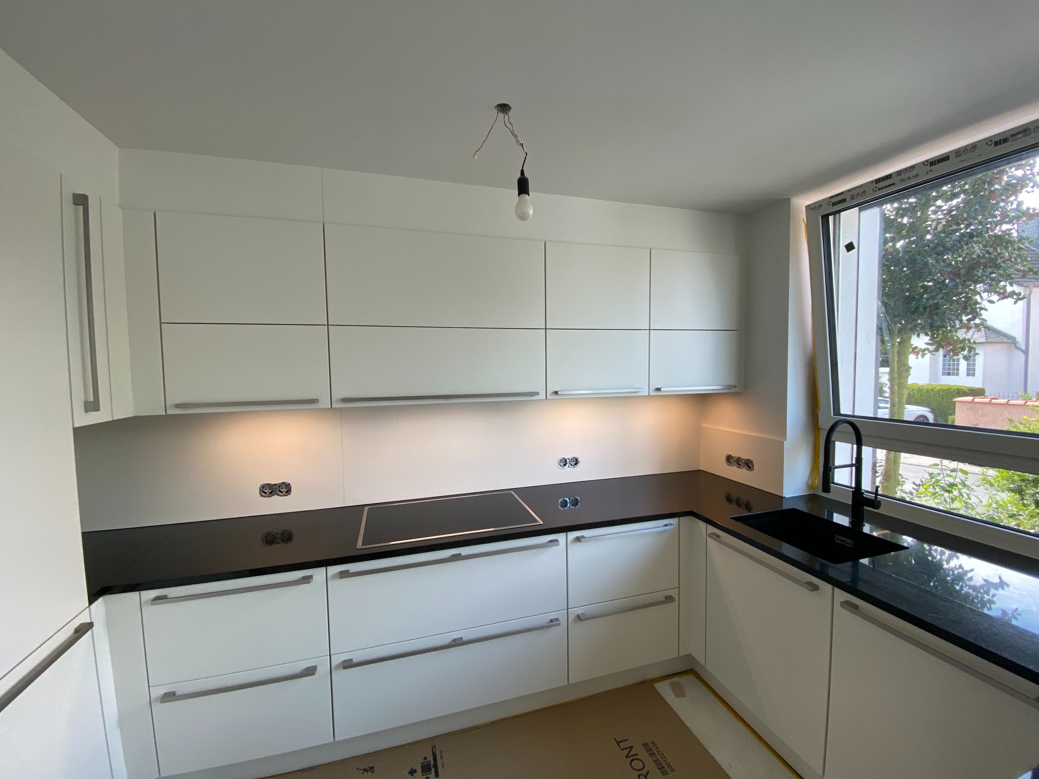 Nowoczesna, biała kuchnia z czarnym blatem i widokiem z okna na zieleń, z surową żarówką zwisającą z sufitu.