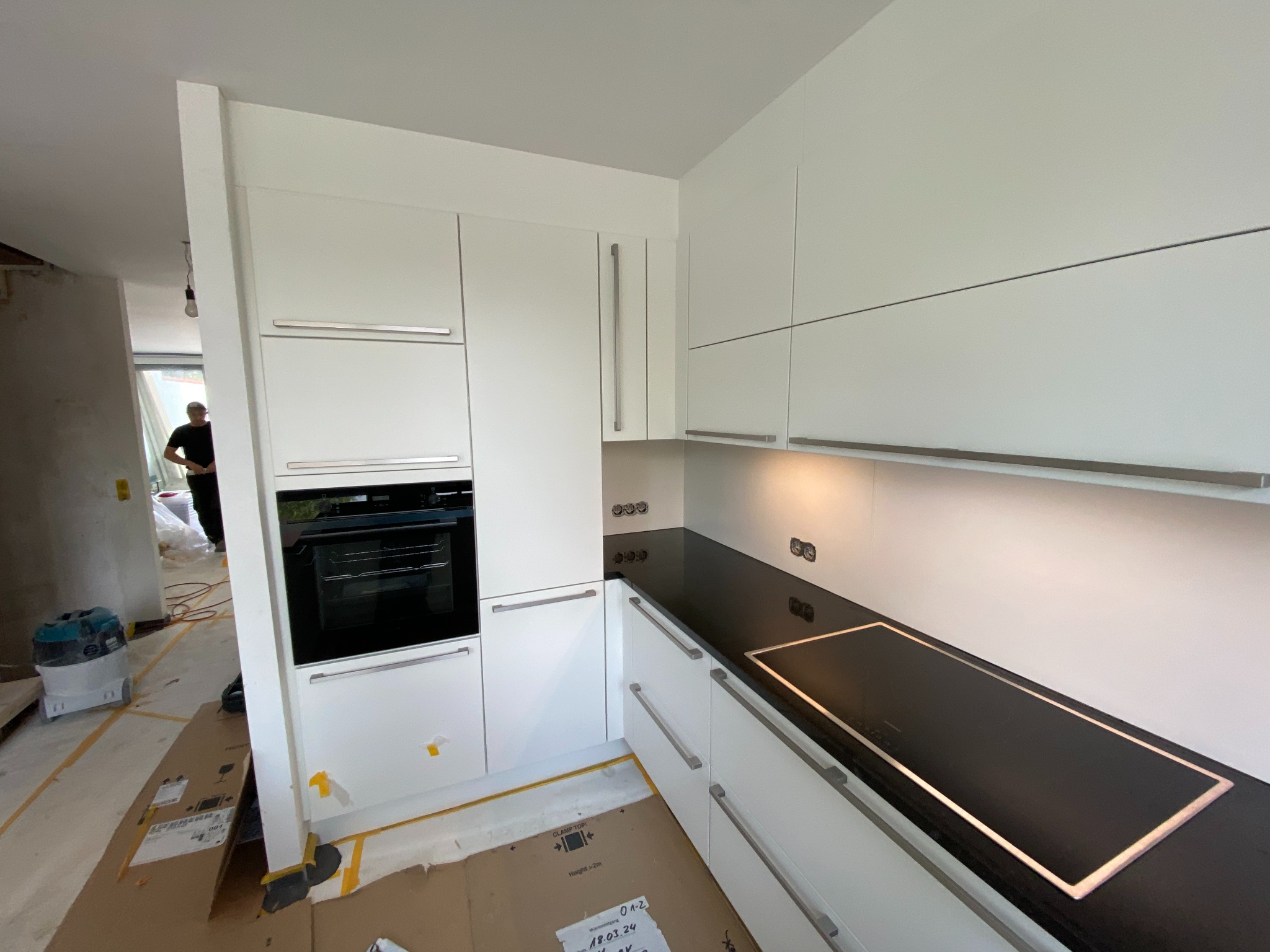 Montaż białej kuchni z czarnym blatem i płytą indukcyjną, widoczne kartony zabezpieczające podłogę i odkurzacz przemysłowy, osoba w tle.