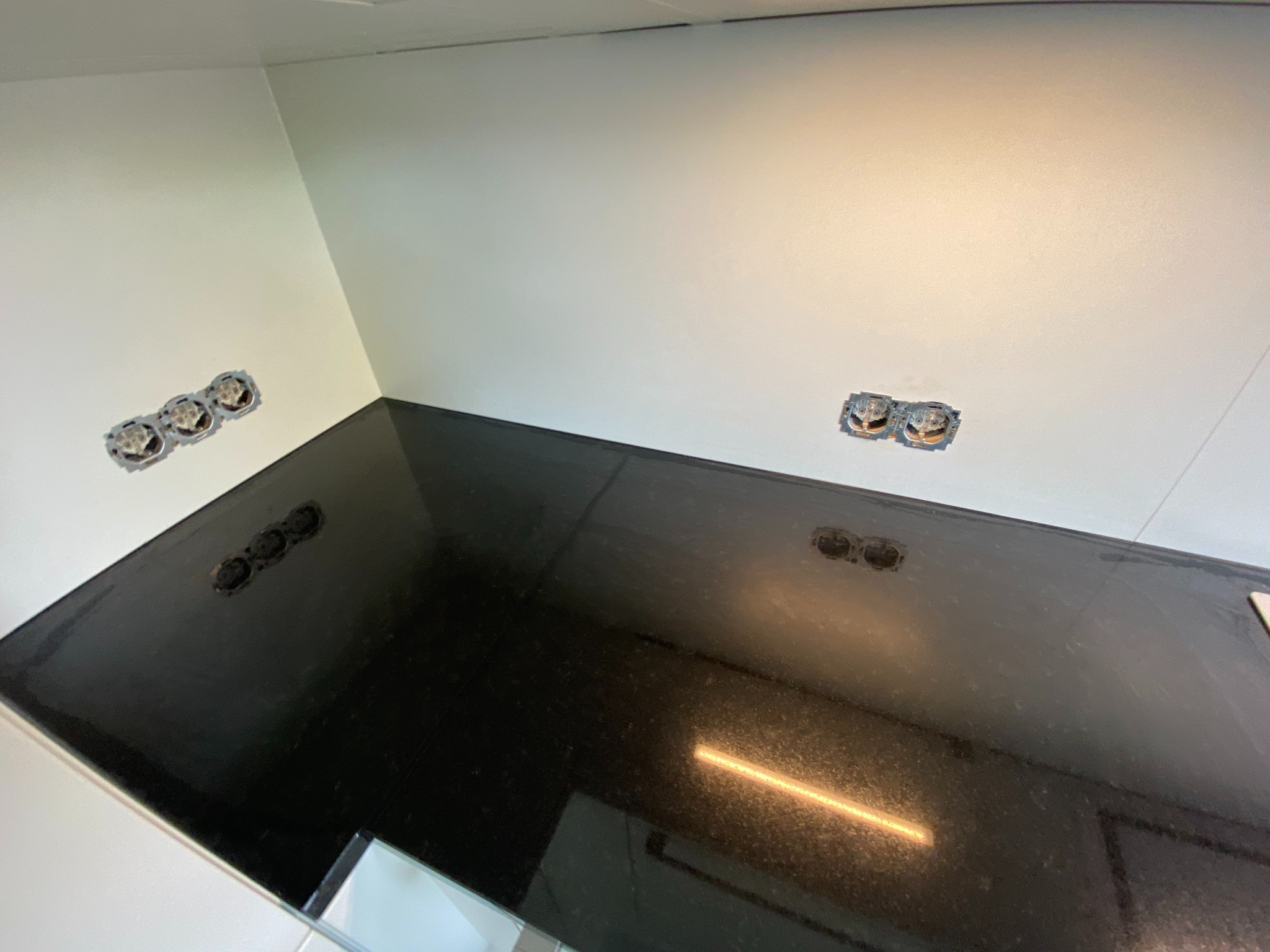 Czarny, błyszczący blat kuchenny z wyciętymi otworami na gniazdka elektryczne, w tle białe ściany z puszkami instalacyjnymi.