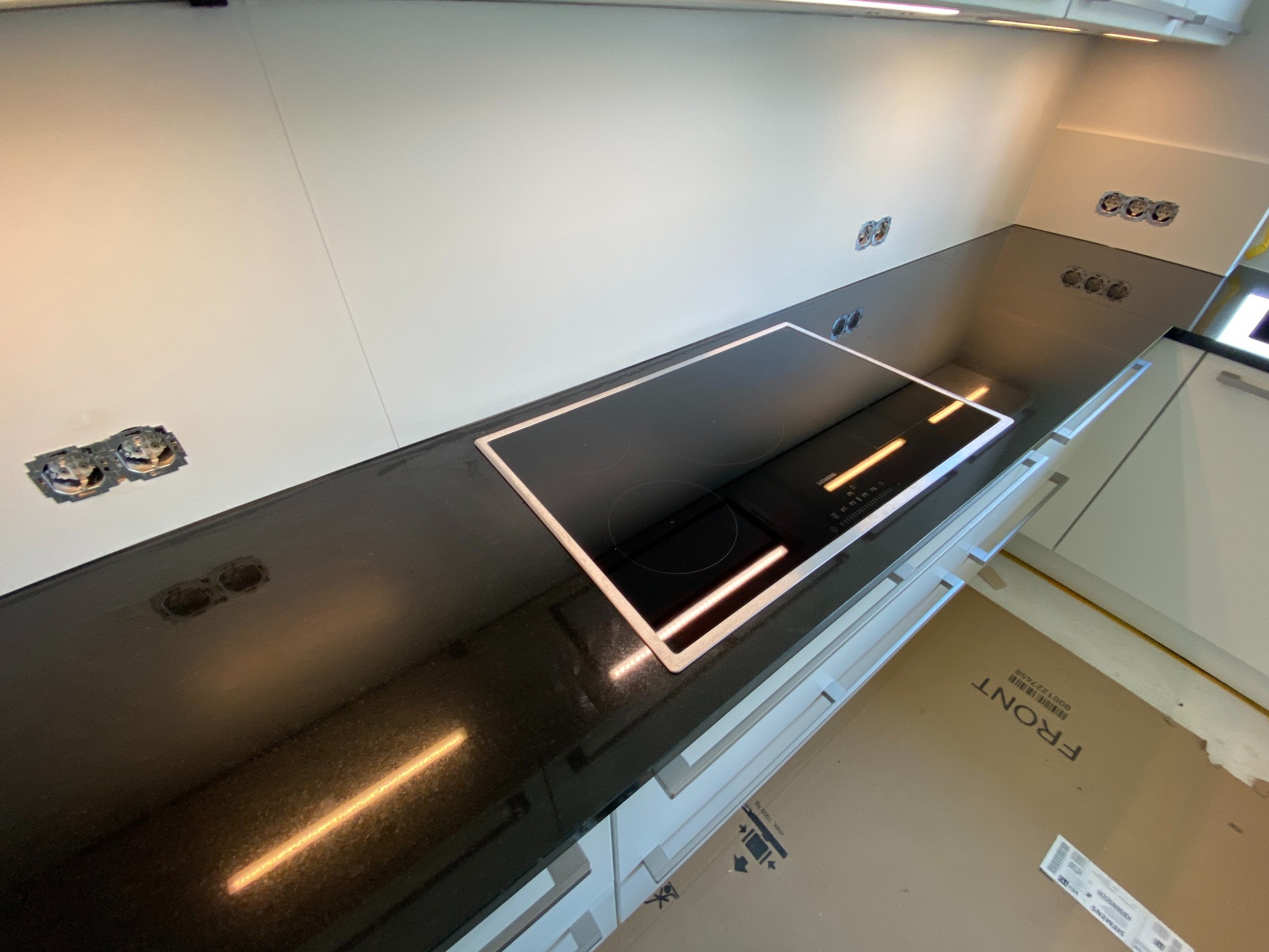 Czarny, błyszczący blat kuchenny z płytą indukcyjną, widoczne puszki elektryczne w ścianie, kartonowe zabezpieczenie pod szafkami.