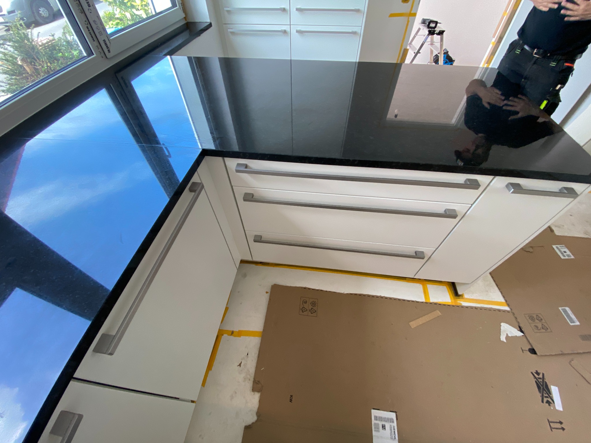 Czarny blat kuchenny z widocznym odbiciem osoby, białe szafki z poziomymi uchwytami, okno z widokiem na zieleń na zewnątrz, podłoga zabezpieczona kartonem.