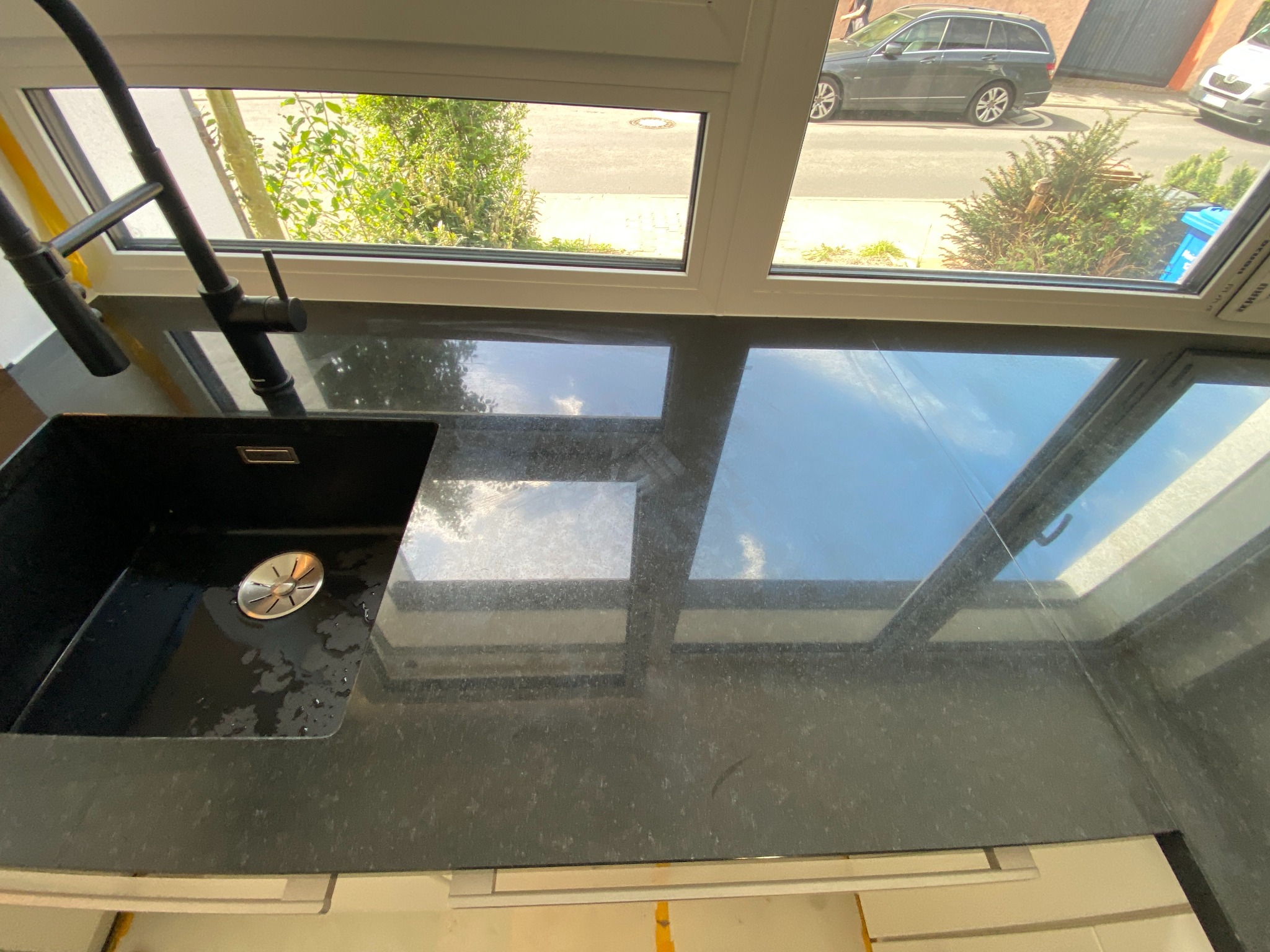 Czarny, granitowy blat kuchenny z wbudowanym, czarnym zlewem i nowoczesną baterią, widok z okna na ulicę z samochodami i zielenią.