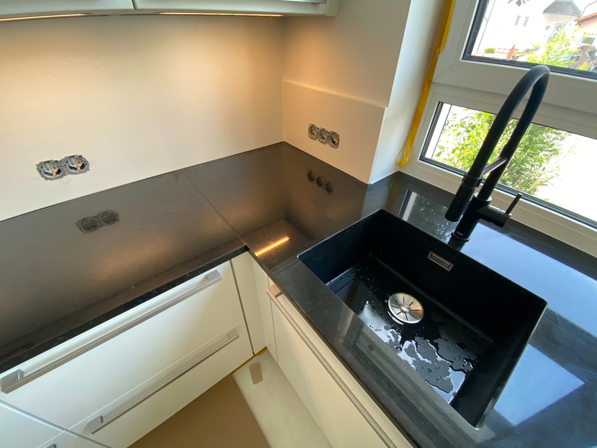 Czarny, granitowy blat kuchenny z prostokątnym zlewem i minimalistyczną, czarną baterią, widoczne gniazdka elektryczne w ścianie i fragment okna z widokiem na zieleń.