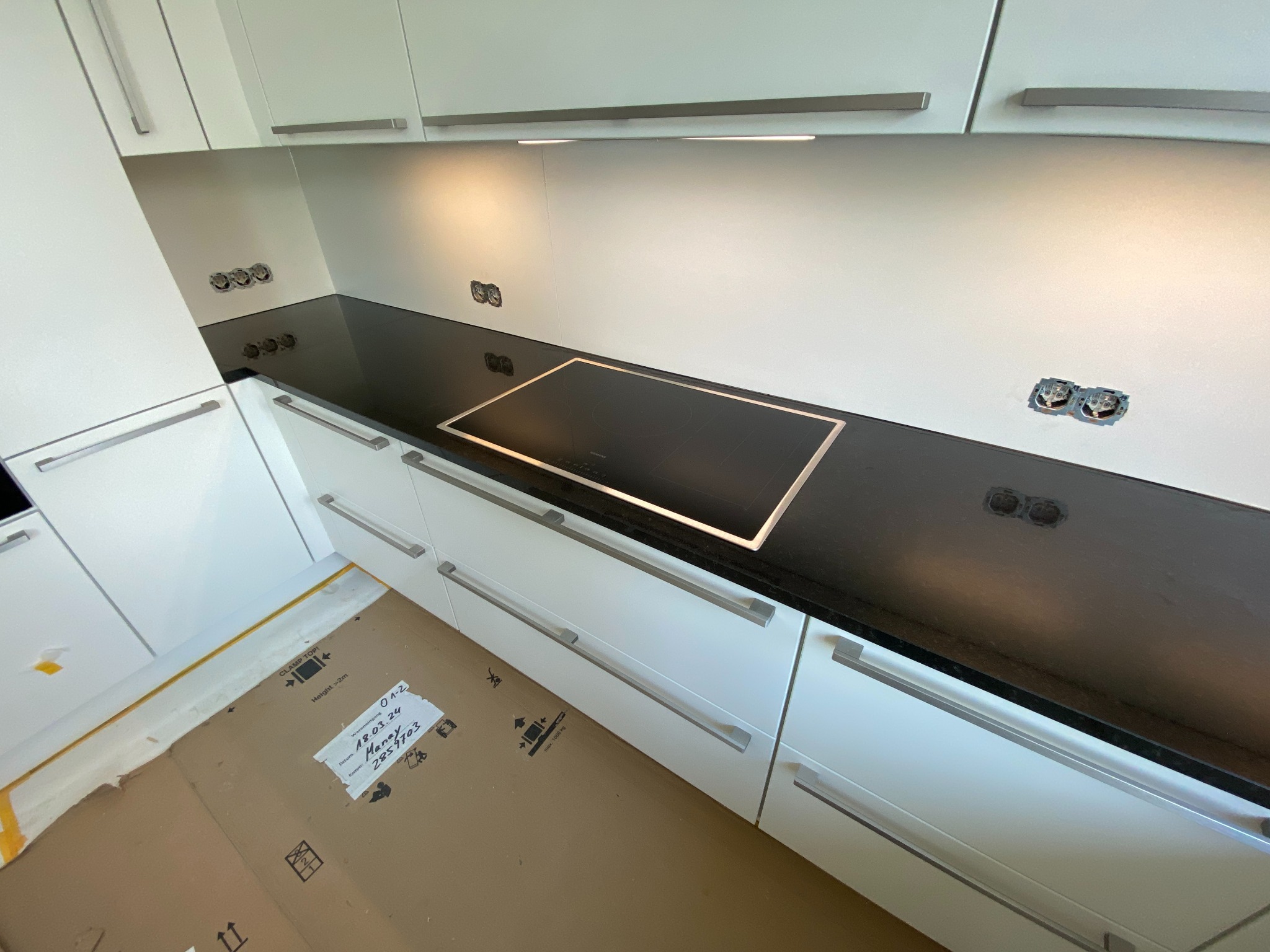 Nowoczesna, biała zabudowa kuchenna z czarnym blatem i płytą indukcyjną; widoczne niezabezpieczone gniazdka elektryczne i kartonowe zabezpieczenie podłogi.