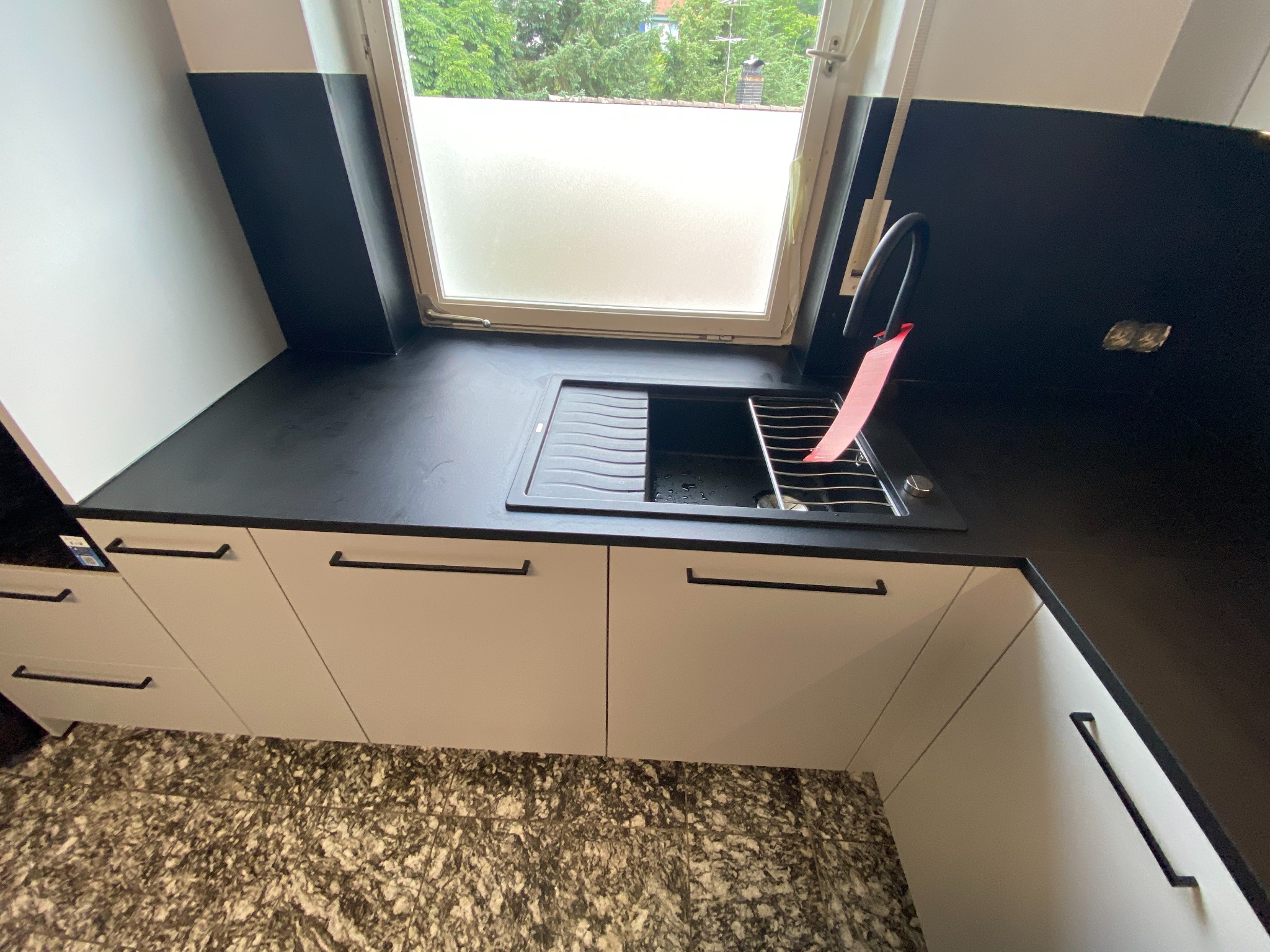 Nowoczesna kuchnia z białymi szafkami z czarnymi uchwytami, czarnym blatem i zlewozmywakiem pod oknem z matową szybą, widok z góry na marmurkową podłogę.