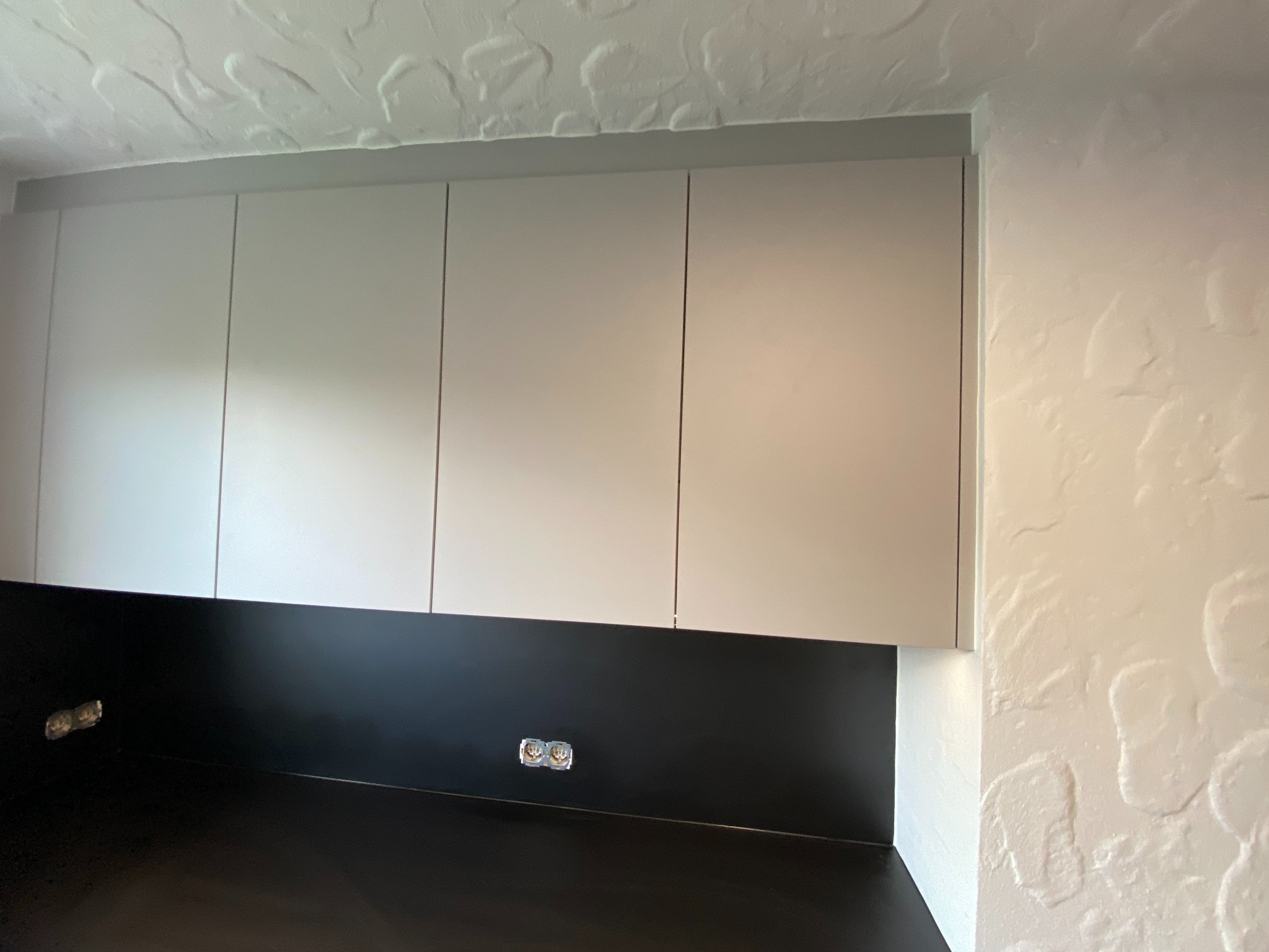 Górne szafki kuchenne w kolorze jasnoszarym, z czarnym panelem nad blatem, widoczne gniazdka elektryczne, ściana z fakturą.