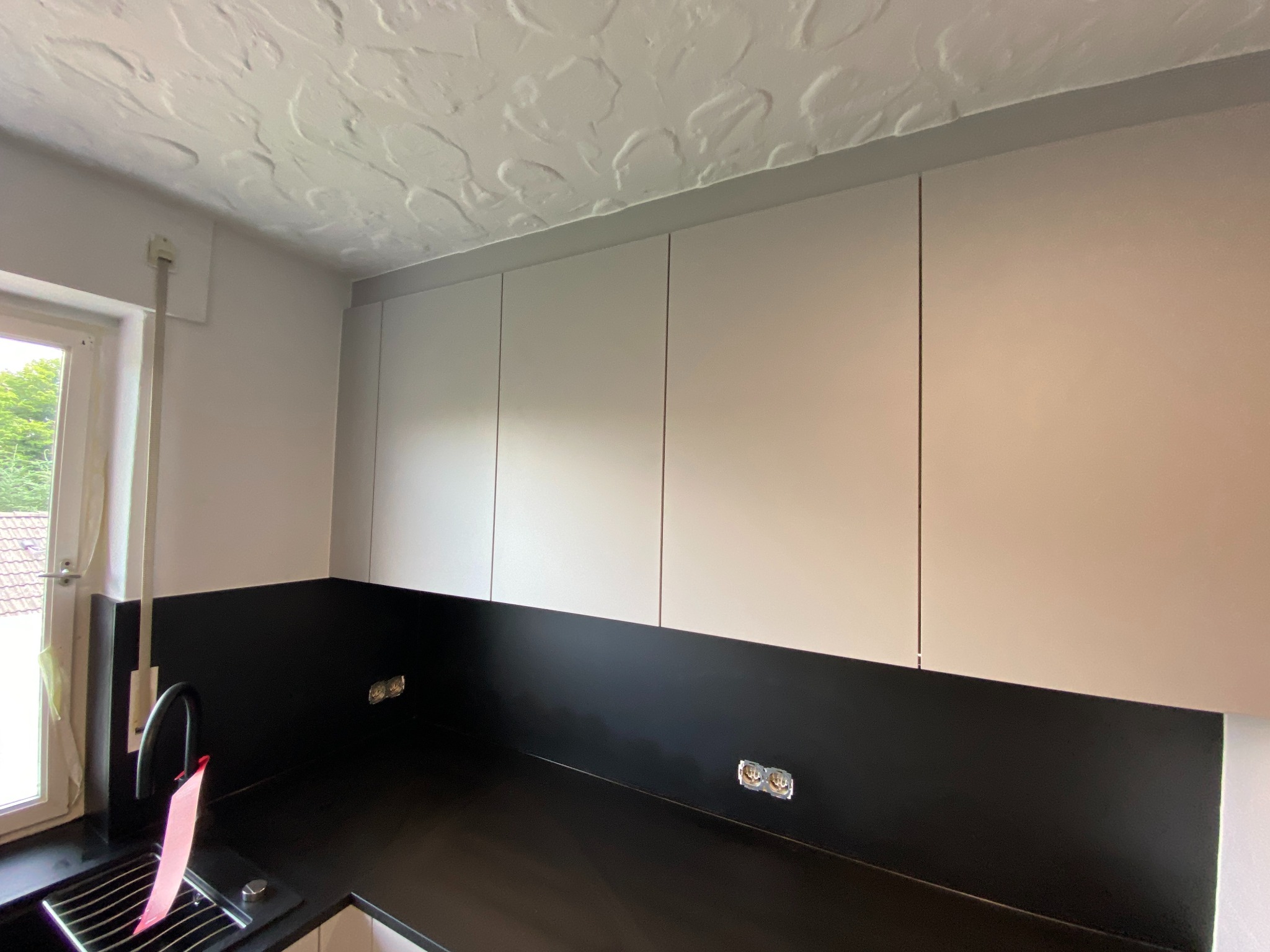 Nowoczesna kuchnia: górne szafki w odcieniach bieli, czarny blat i panel ścienny, widoczne gniazdka elektryczne, faktura sufitu.