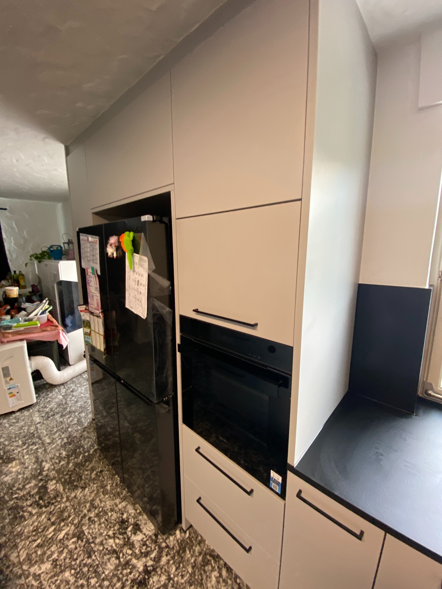 Nowoczesna kuchnia z jasnymi frontami szafek, czarnym piekarnikiem i lodówką, widok z boku, na blacie czarny panel.