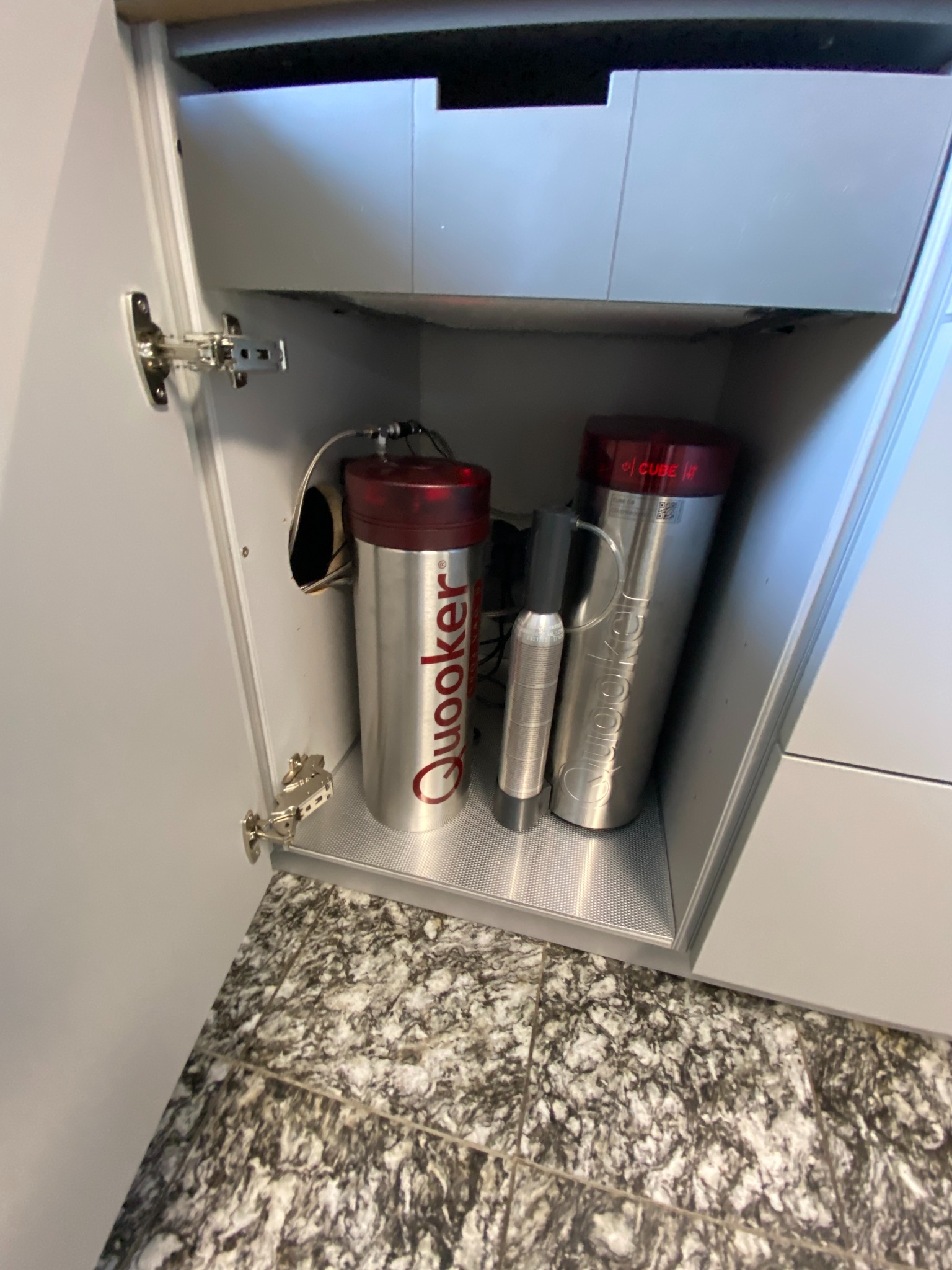 Wnętrze szafki kuchennej z systemem Quooker Cube do filtrowania i gazowania wody, widoczne srebrne zbiorniki z czerwonymi elementami, zainstalowane w szafce z szarą półką i marmurowym blatem.