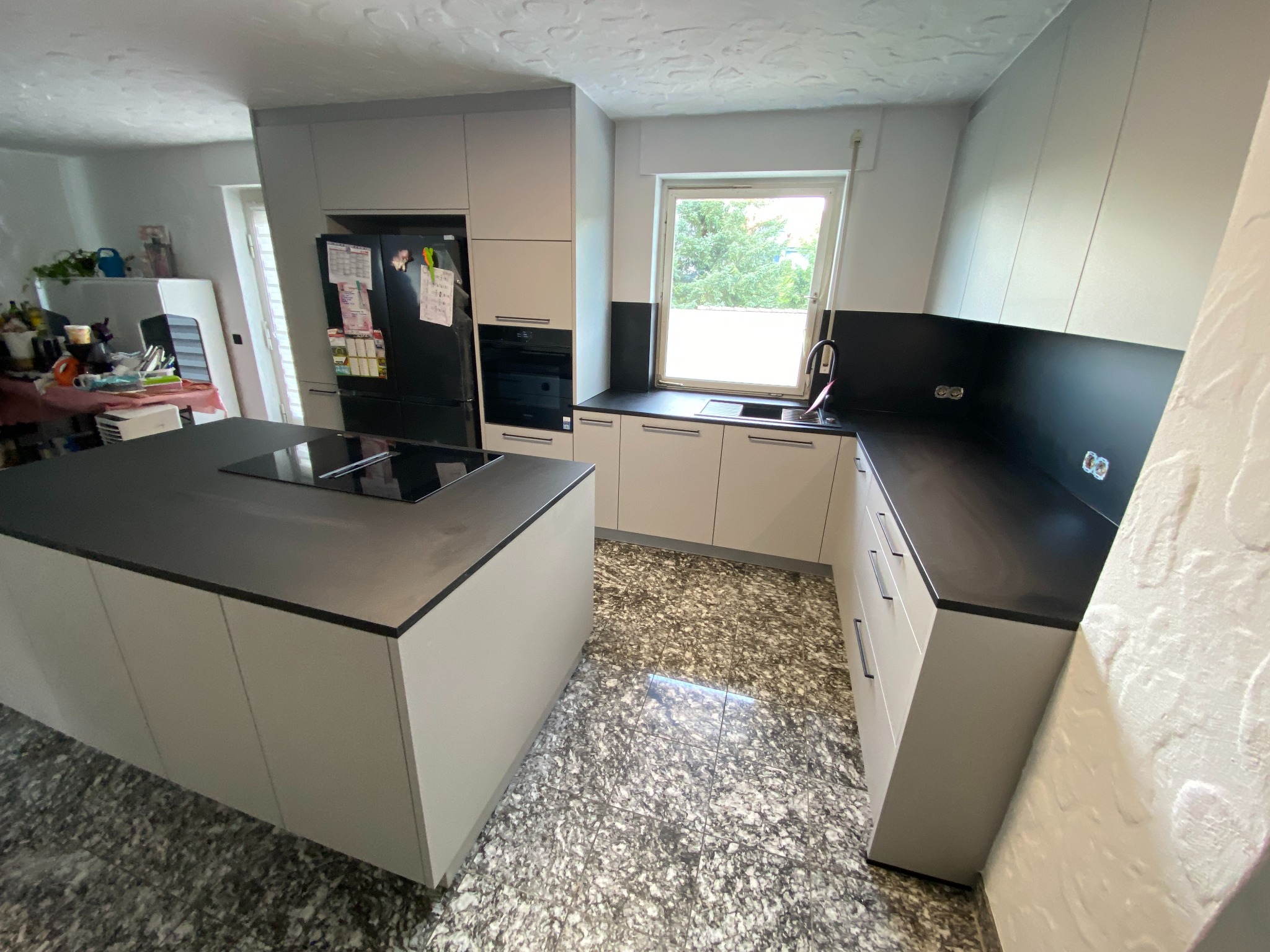 Nowoczesna kuchnia z wyspą, jasne szafki, czarny blat, podłoga w marmurkowy wzór, okno z widokiem na zieleń.