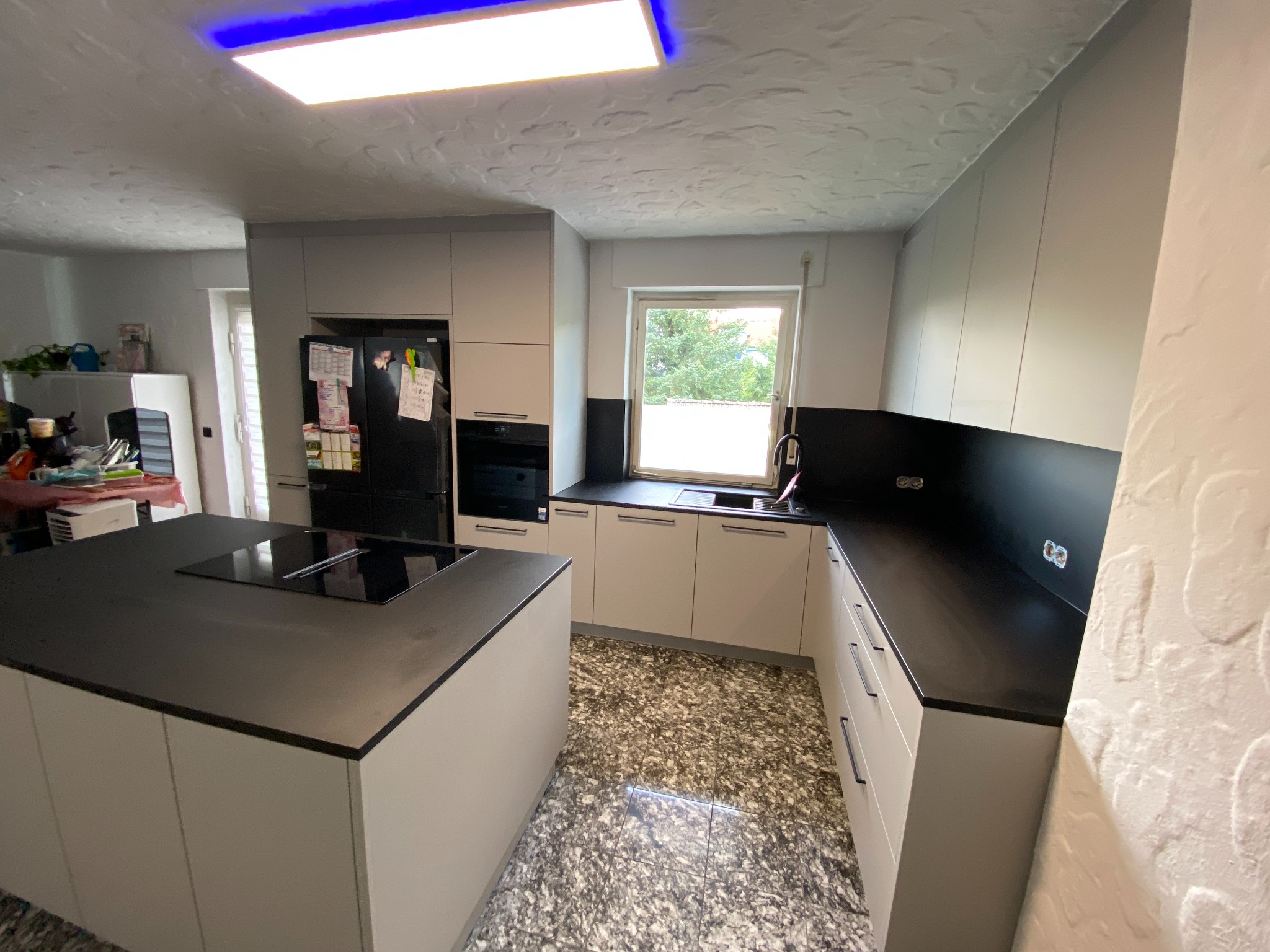 Nowoczesna kuchnia z wyspą i czarnymi blatami, jasne szafki bez uchwytów, wzorzysta podłoga, oświetlenie sufitowe LED.