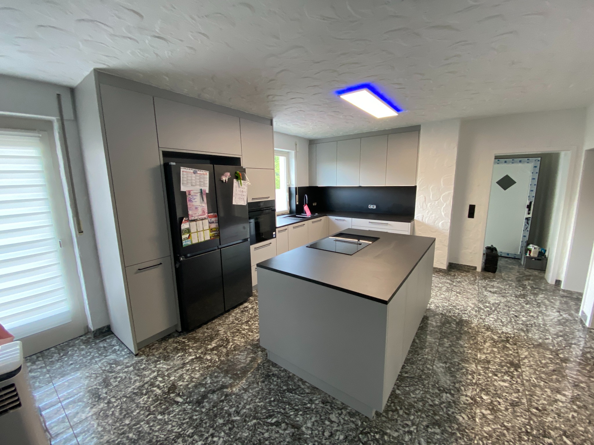 Nowoczesna kuchnia w szarościach z wyspą kuchenną i czarną lodówką, oświetlona lampą z niebieską poświatą, na podłodze wzorzyste płytki.