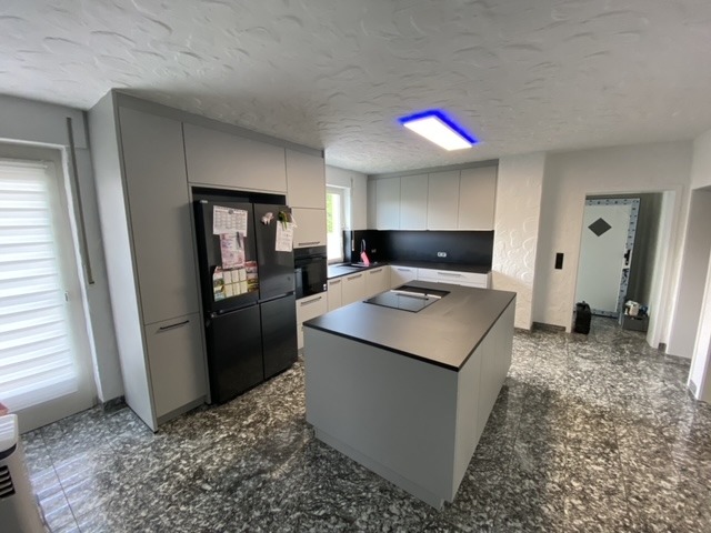 Nowoczesna kuchnia w szarościach z wyspą i czarnym sprzętem AGD, widok na nieregularną podłogę i drzwi wewnętrzne.