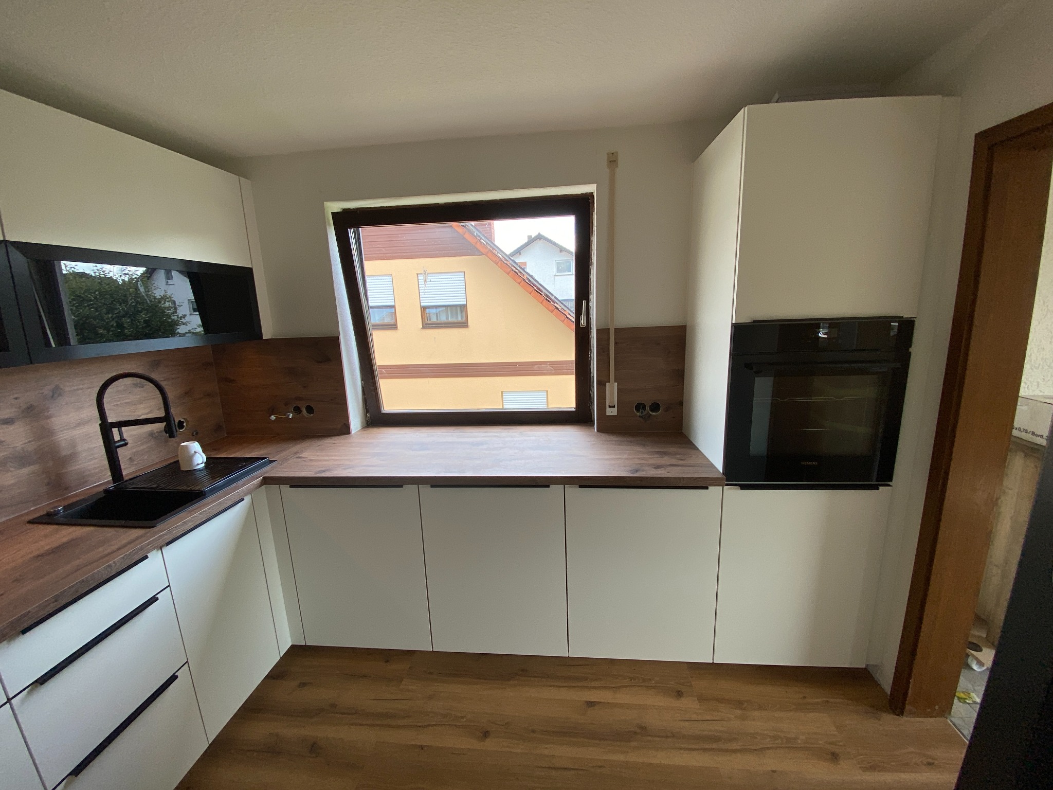 Nowoczesna kuchnia z białymi frontami szafek i blatem imitującym drewno, czarny zlewozmywak i bateria, okno z widokiem na budynek mieszkalny.