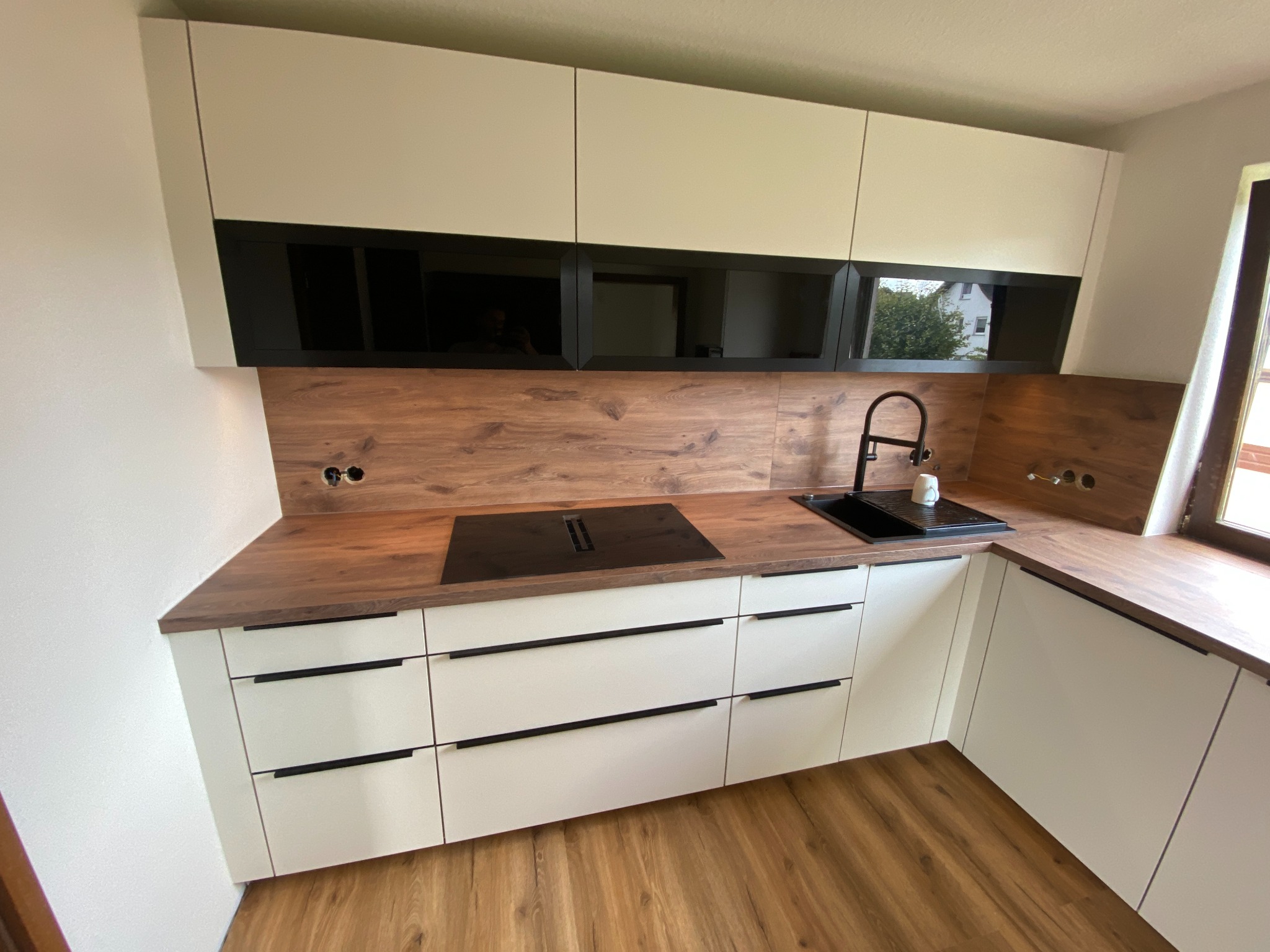 Nowoczesna kuchnia z białymi szafkami dolnymi i górnymi z czarnymi, szklanymi frontami, drewnianym blatem i panelem ściennym, czarną armaturą i płytą indukcyjną.