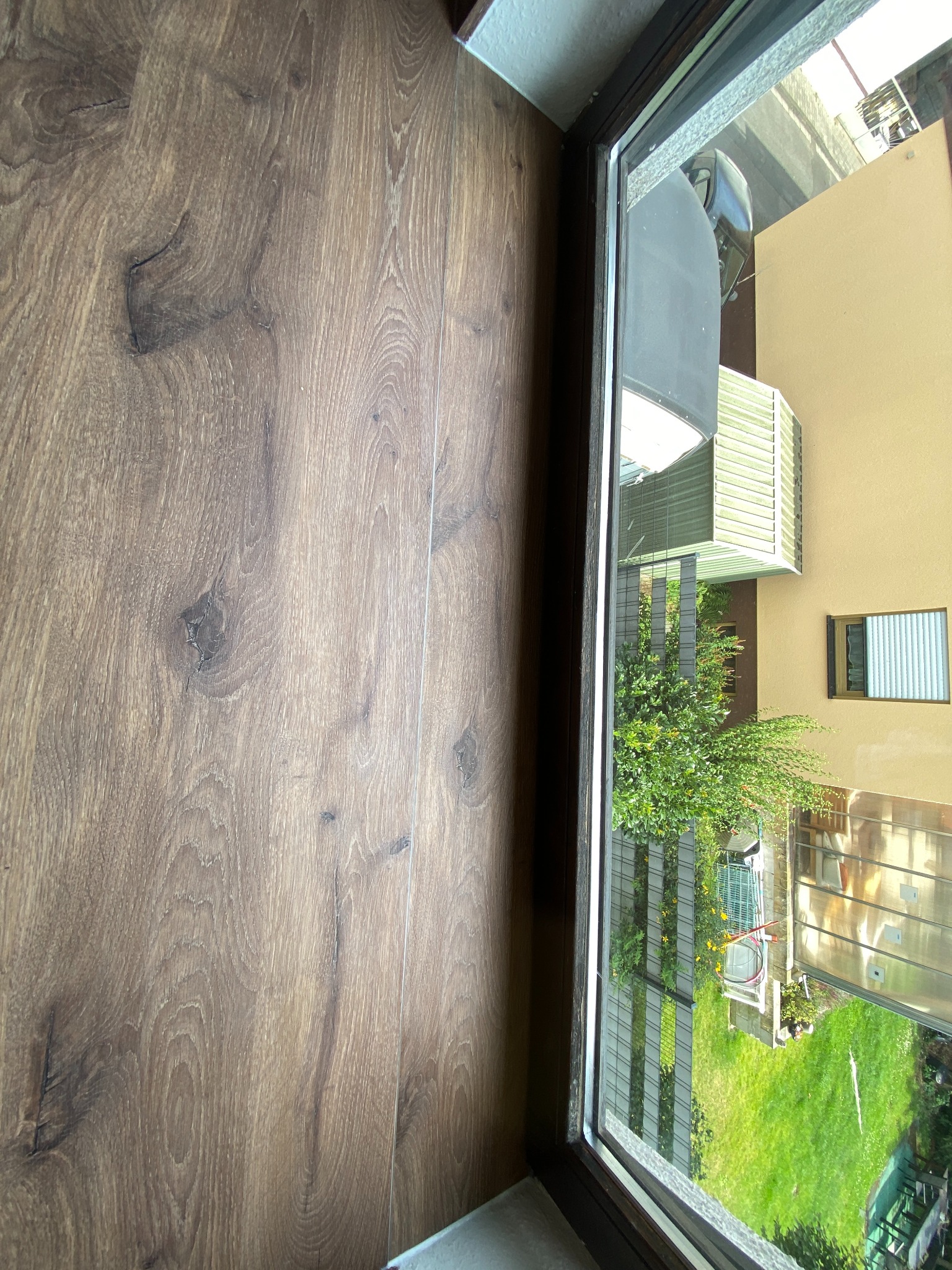 Fragment drewnianej podłogi w odcieniach brązu, widoczna przez okno z widokiem na zielony ogród i budynek z jasną elewacją.