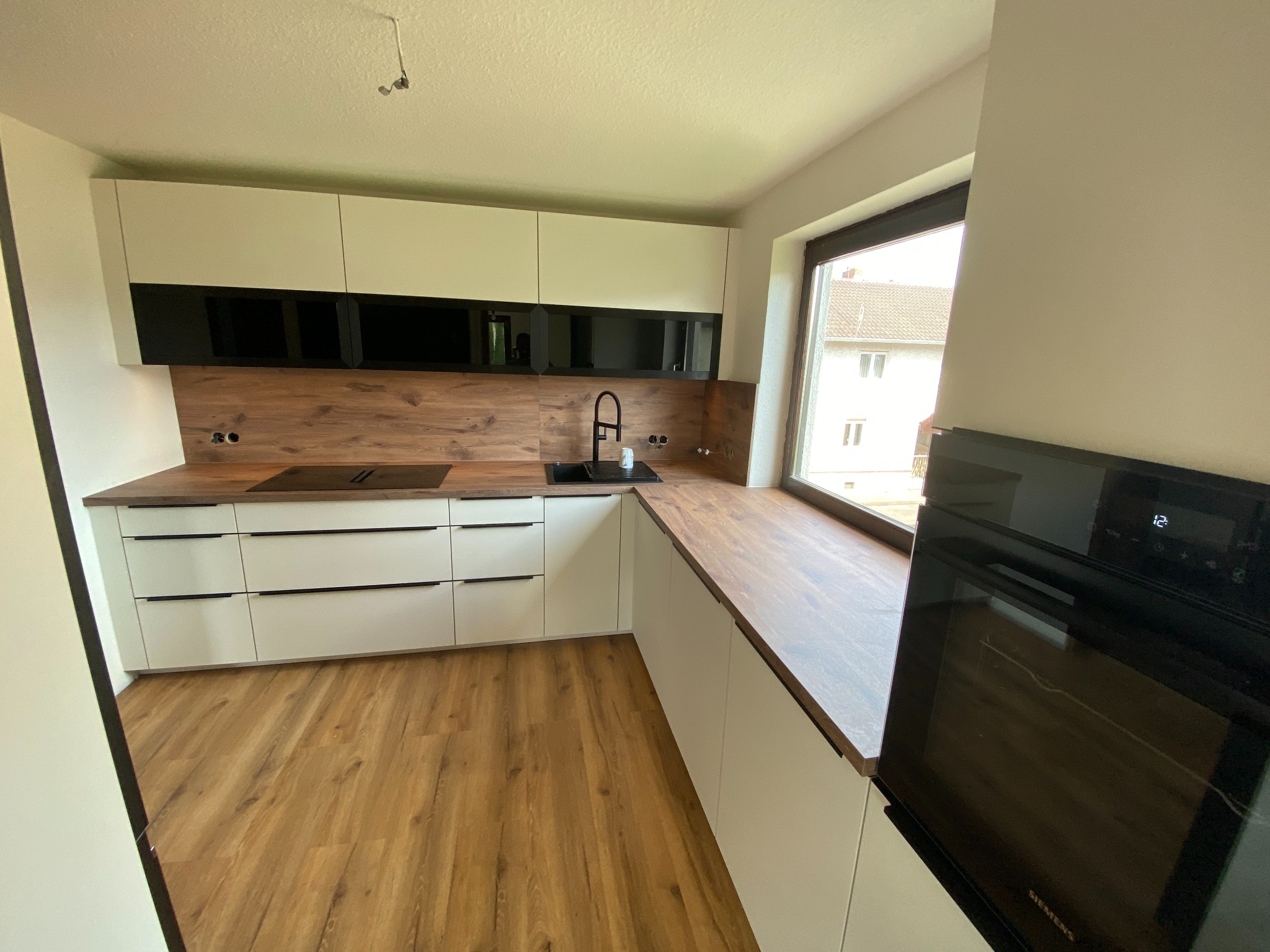 Nowoczesna kuchnia z białymi szafkami, czarnymi akcentami i drewnianym blatem roboczym, widok z boku na piekarnik Siemens, jasna podłoga i okno z widokiem na dom.