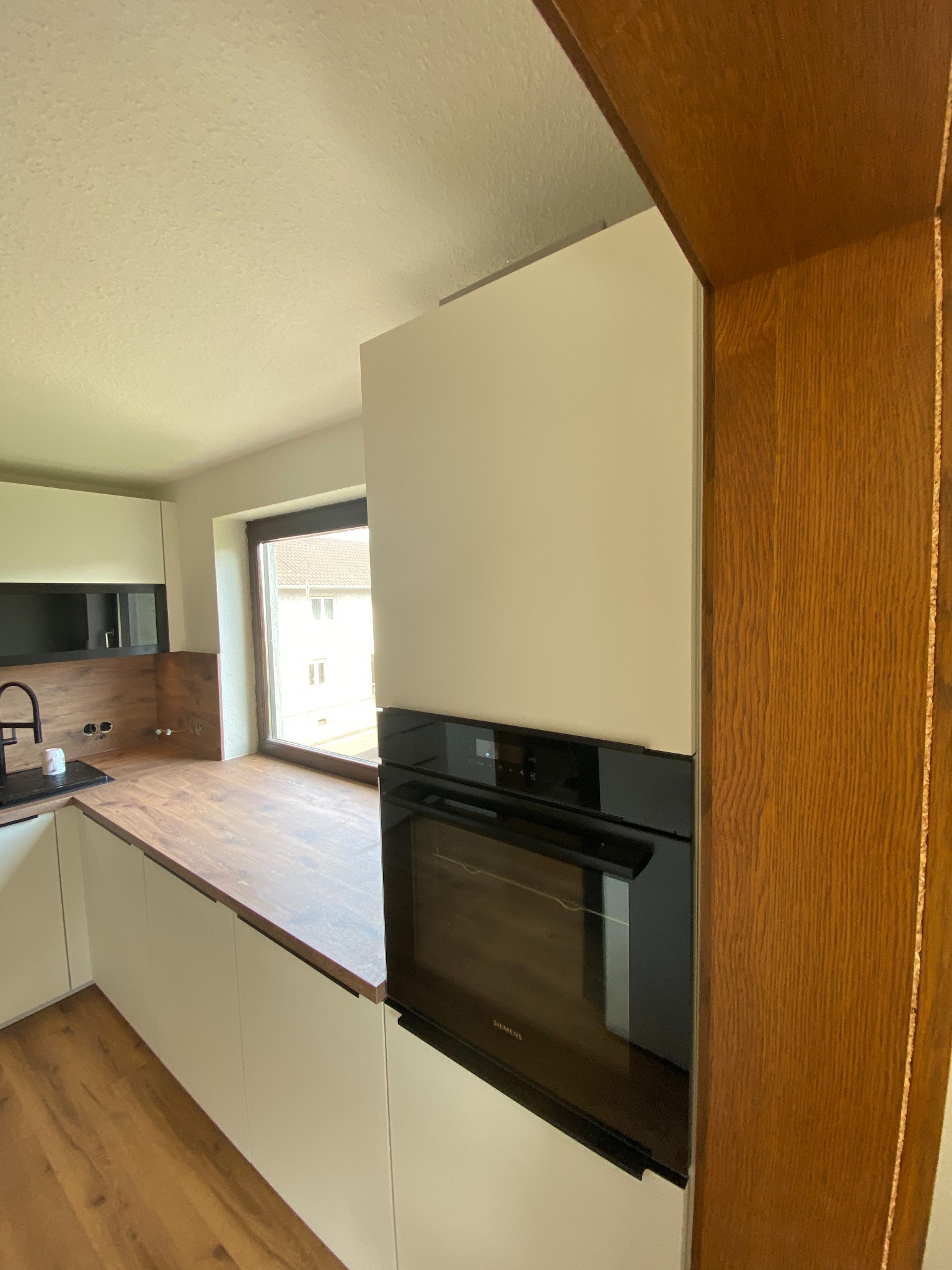 Nowoczesna kuchnia z białymi frontami, drewnianym blatem i zabudowanym piekarnikiem Siemens, widok z perspektywy wnętrza, z oknem w tle.