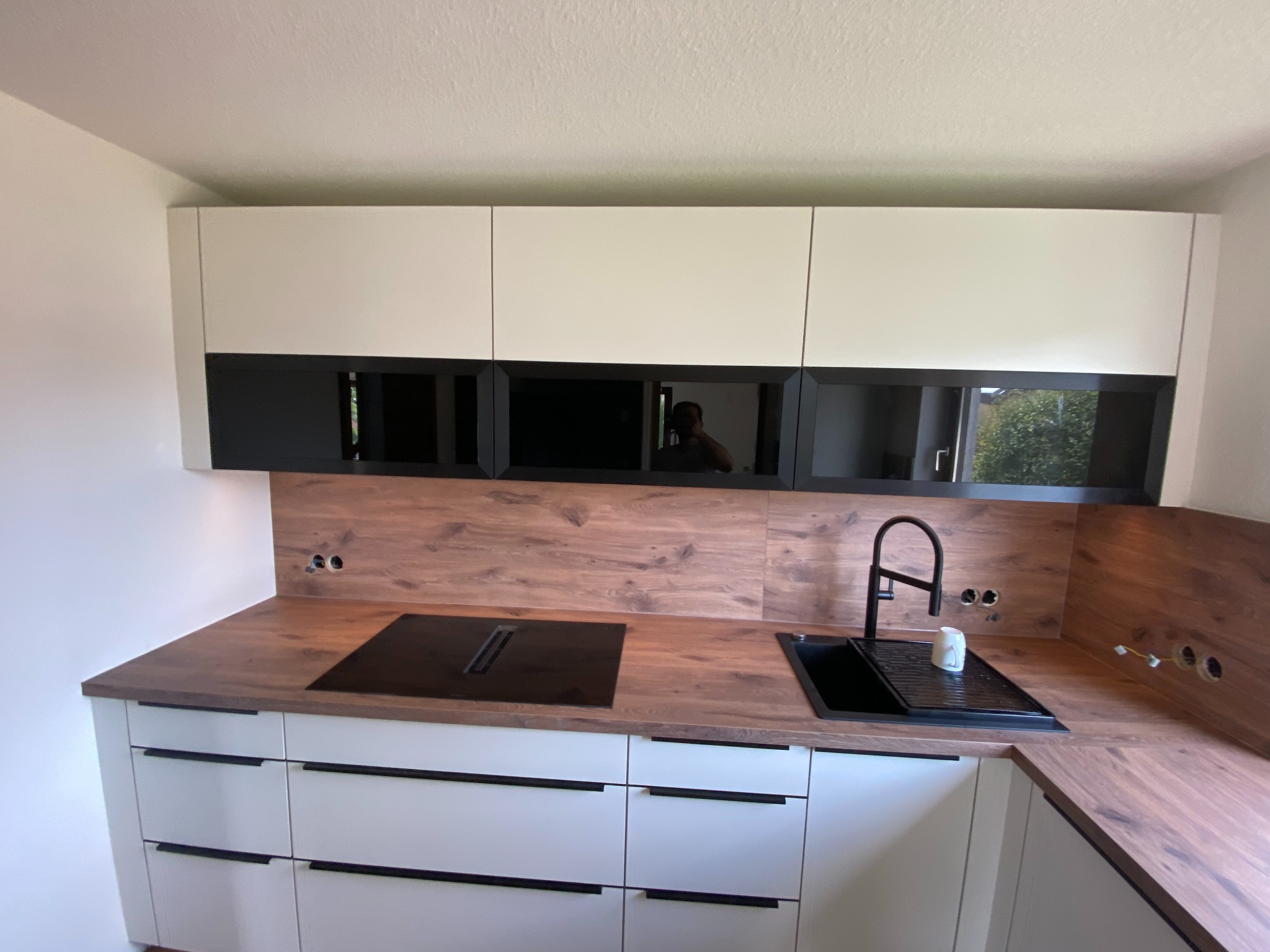 Nowoczesna kuchnia z białymi szafkami z czarnymi uchwytami, drewnianym blatem i panelem ściennym. W tle widoczne odbicie osoby w szklanych frontach szafek wiszących.