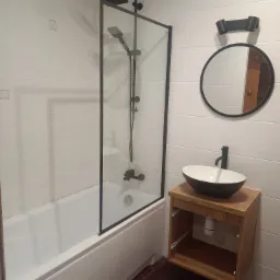 Nowoczesna łazienka z kabiną prysznicową typu walk-in z czarną ramą, owalną umywalką nablatową na drewnianej szafce i okrągłym lustrem z czarną obwódką na białych kafelkach.