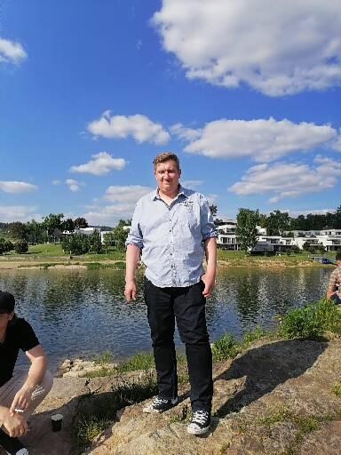 Mężczyzna w koszuli i czarnych spodniach stoi na tle jeziora i błękitnego nieba z chmurami.