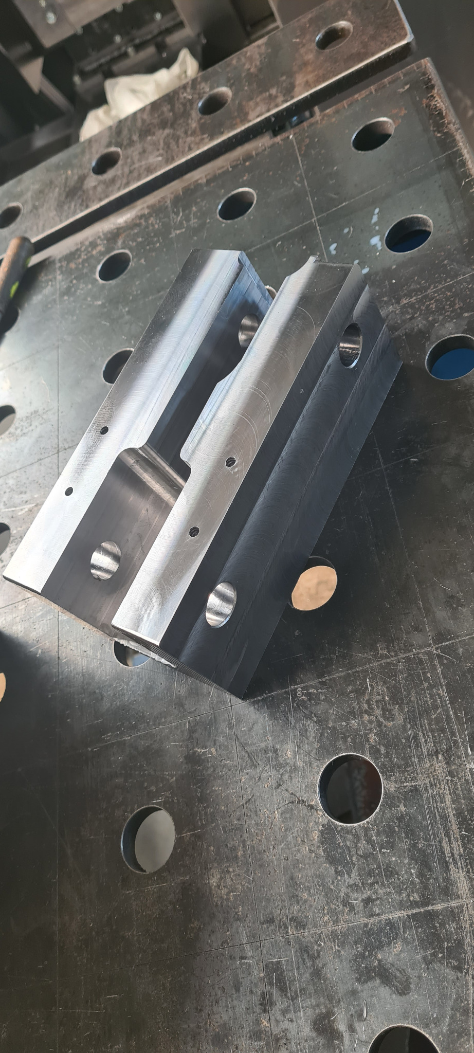 Detal z metalu z otworami, obrobiony na maszynie CNC, leżący na perforowanym stole warsztatowym.