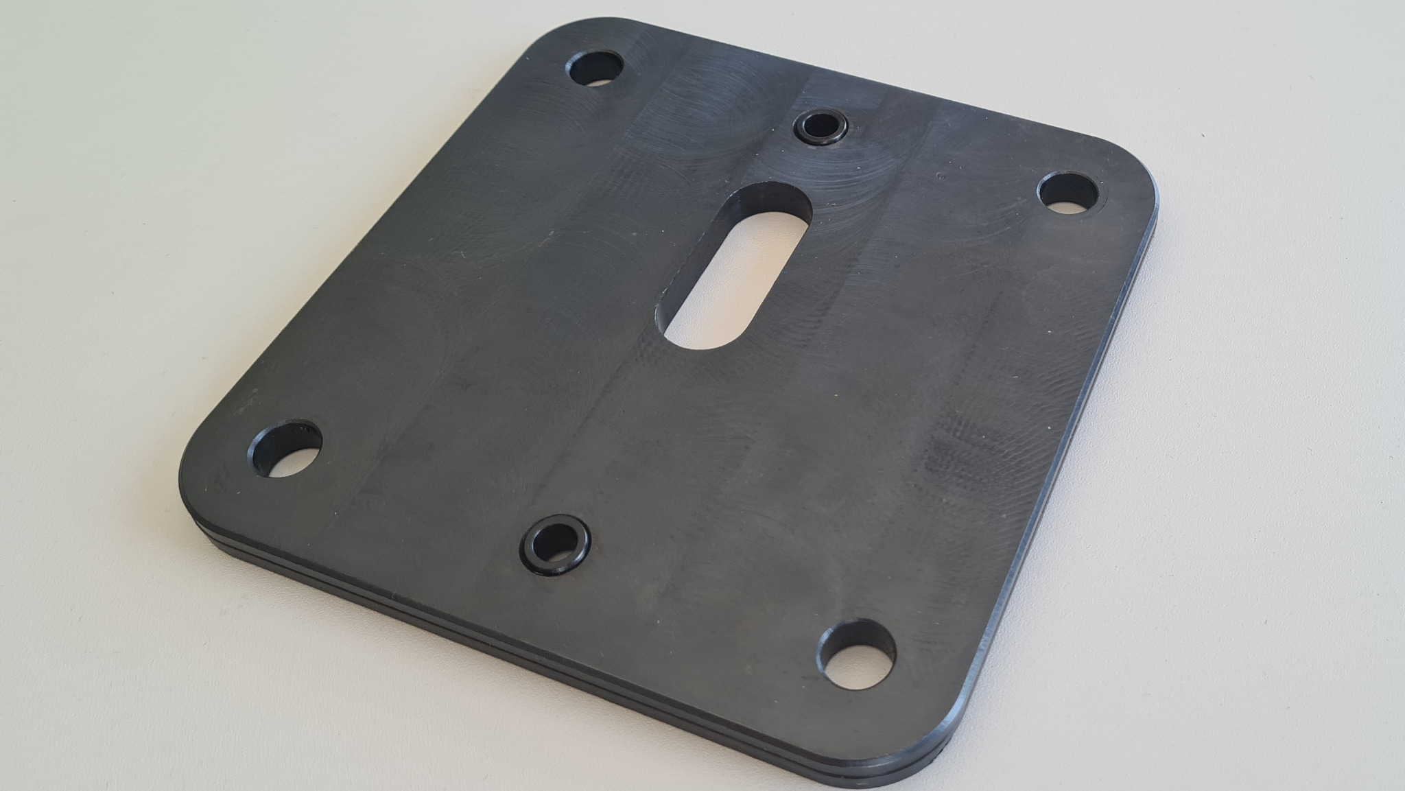 Czarna, kwadratowa płyta z tworzywa sztucznego po obróbce CNC, z zaokrąglonymi rogami, czterema otworami przelotowymi, dwoma otworami gwintowanymi i podłużnym otworem centralnym, leżąca na jasnym tle.