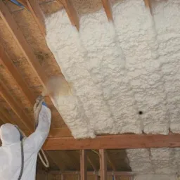 Pracownik w kombinezonie ochronnym aplikuje piankę izolacyjną na poddaszu, widoczna drewniana konstrukcja dachu.