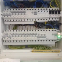 Rozdzielnica elektryczna z zainstalowanymi bezpiecznikami, przewodami w różnych kolorach i listwami zaciskowymi, widoczne zielone diody LED sygnalizujące działanie, dolna część zdjęcia jest lekko...