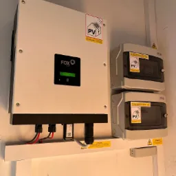 Biały falownik marki FoxESS z wyświetlaczem pokazującym aktualną produkcję energii, zamontowany na ścianie obok dwóch rozdzielnic PV-DC i PV-AC z naklejkami ostrzegawczymi.