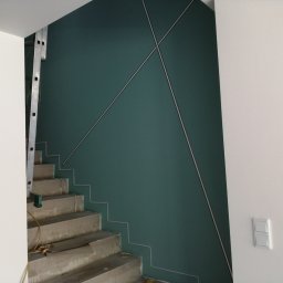 Sufitex - Klatka schodowa w trakcie wykańczania, ściana pomalowana na ciemnozielono z geometrycznym wzorem z listew, surowe betonowe schody zabezpieczone folią, widoczna drabina i instalacja elektryczna.