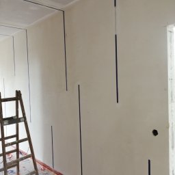 Sufitex - Ściana przygotowana do położenia gładzi, z pionowymi paskami taśmy malarskiej wyznaczającymi miejsca na listwy, drewniana drabina oparta o ścianę, podłoga zabezpieczona folią malarską, widoczne...