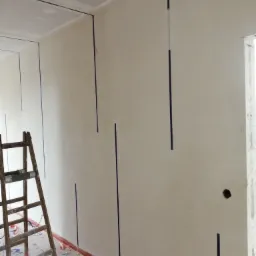 Ściana przygotowana do położenia gładzi, z pionowymi paskami taśmy malarskiej wyznaczającymi miejsca na listwy, drewniana drabina oparta o ścianę, podłoga zabezpieczona folią malarską, widoczne...