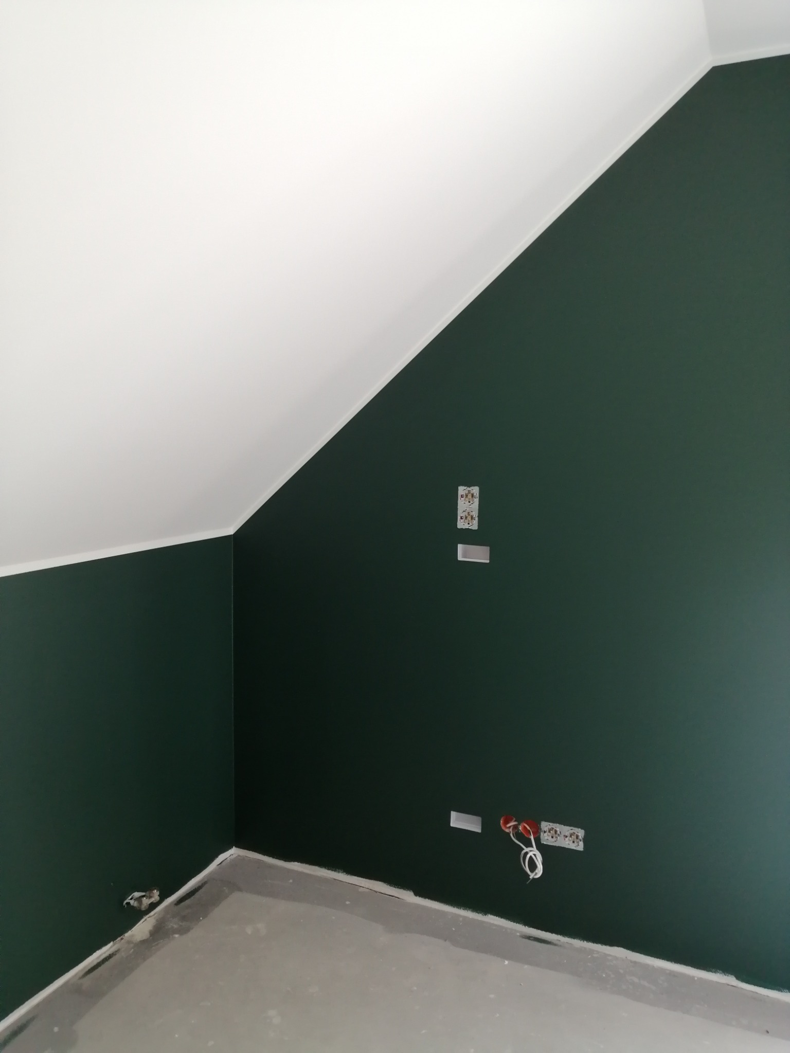 Poddasze po remoncie: ściany pomalowane na ciemnozielony kolor, biały sufit ze skosem, widoczne gniazdka elektryczne i kable wystające ze ściany, surowa podłoga.