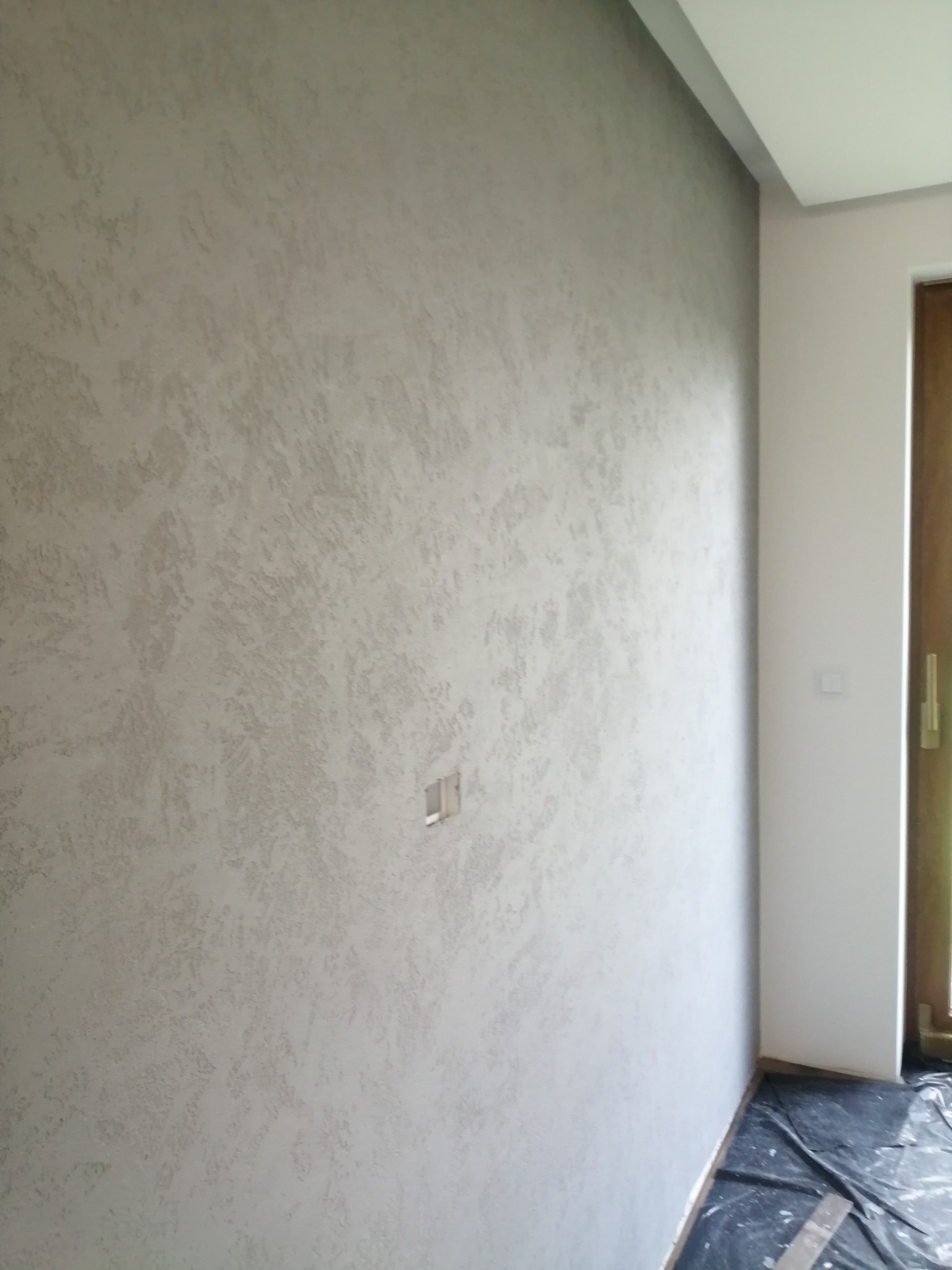 Ściana w pokoju wykończona dekoracyjną strukturą w odcieniach szarości, zabezpieczona folią podłoga, widoczny fragment drzwi.